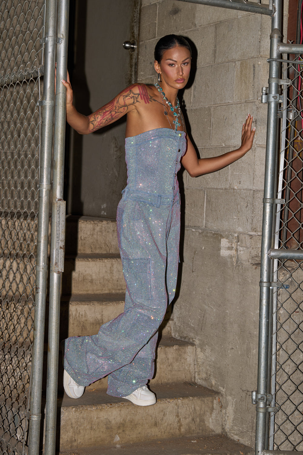 A woman with long dark hair and tattoos poses on concrete steps between chain-link fences, wearing the TOO FLY RHINESTONE DENIM JUMPSUIT and white sneakers against a gray cinderblock wall. 3