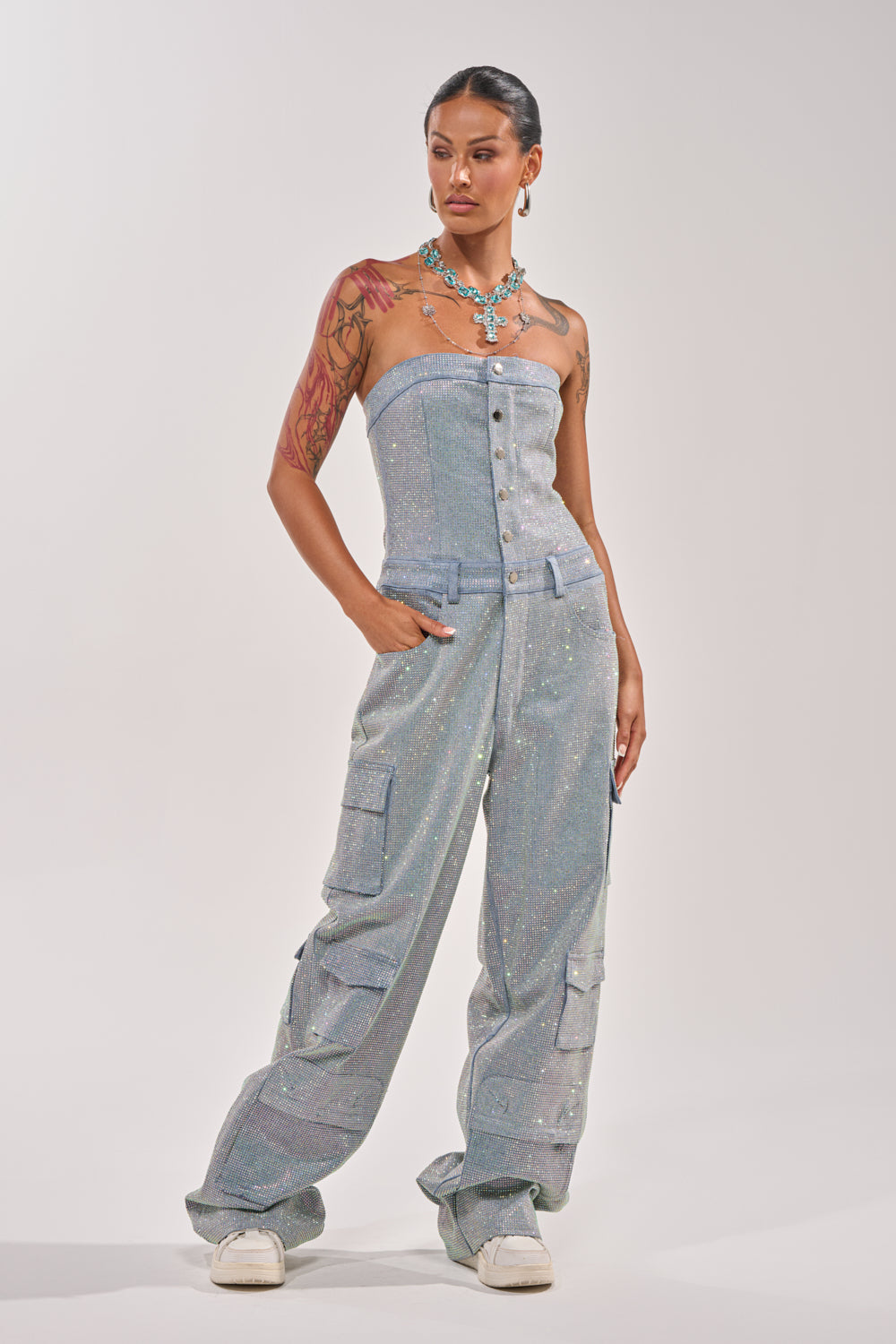 A tattooed woman poses in the TOO FLY RHINESTONE DENIM JUMPSUIT—strapless, button-up, with cargo pockets—styled with a statement necklace and white sneakers against a plain white background. 4