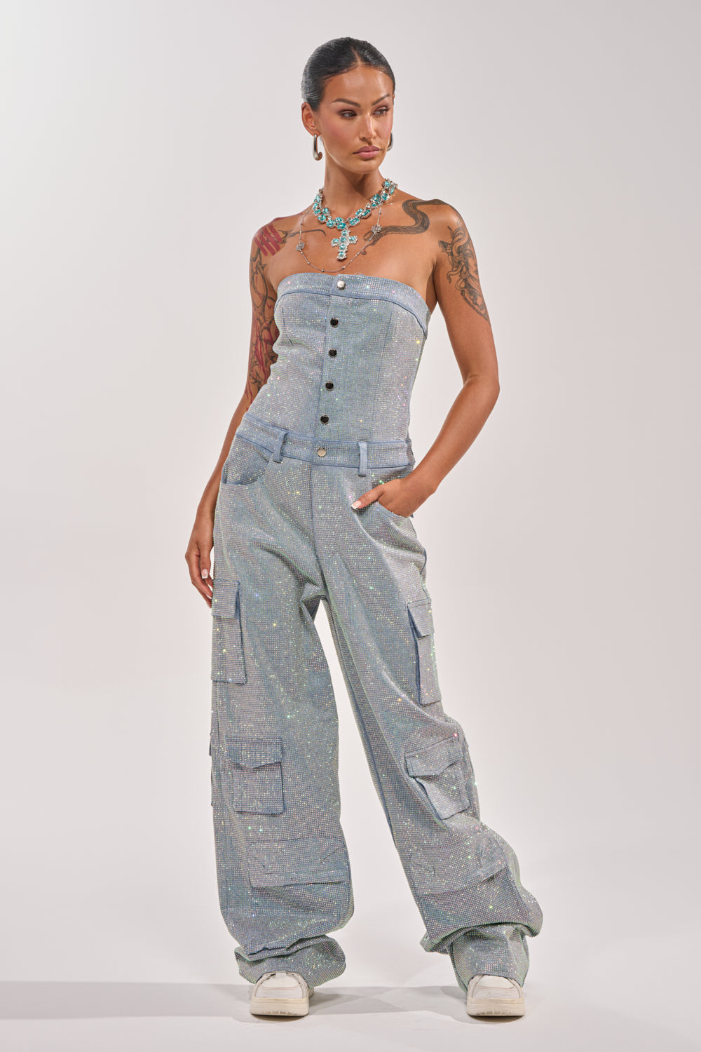 A woman with tattoos stands confidently in the TOO FLY RHINESTONE DENIM JUMPSUIT— a strapless, wide-leg cargo style with subtle sparkles—paired with white sneakers and a turquoise statement necklace against a plain background. 5