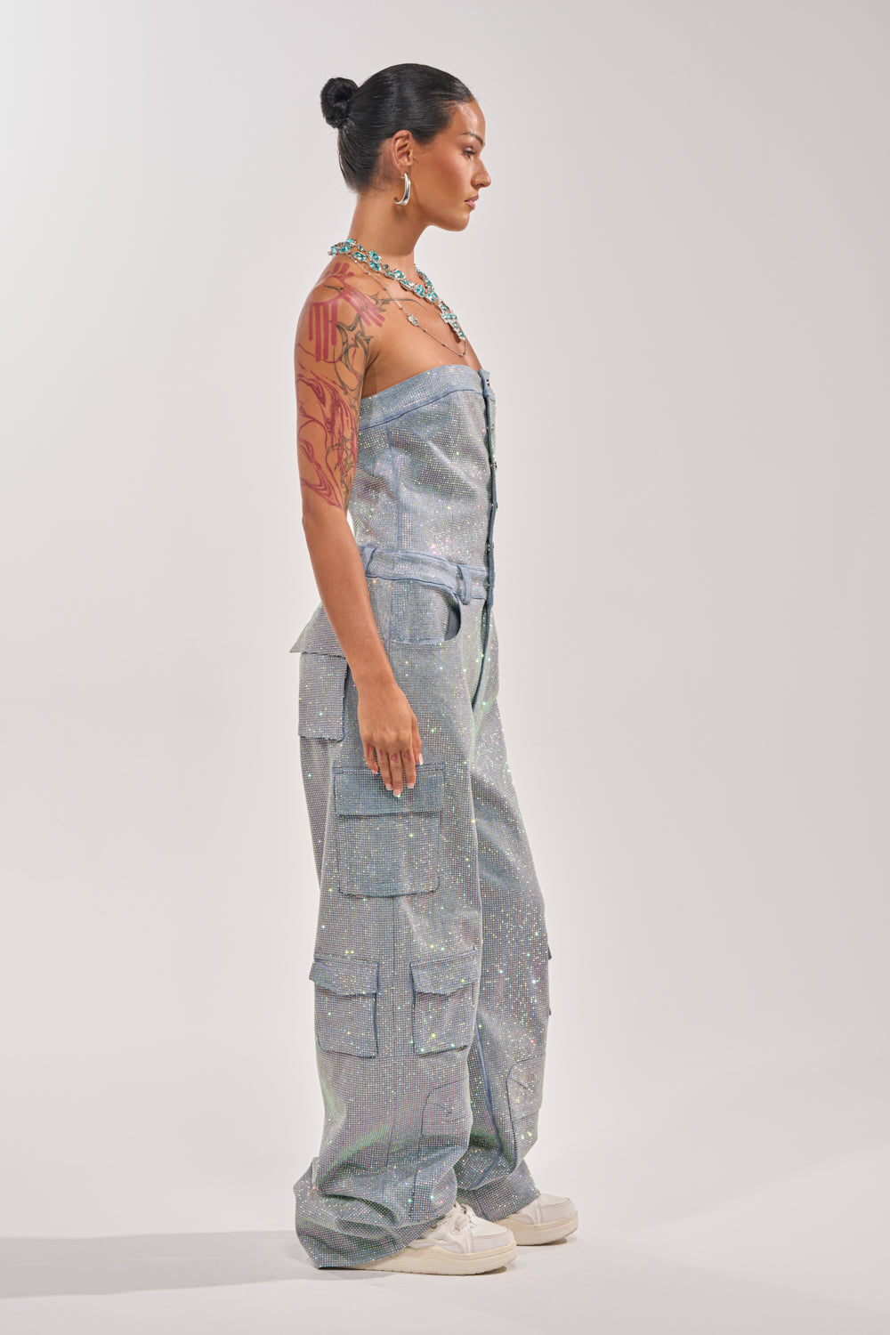 A woman in profile wears the TOO FLY RHINESTONE DENIM JUMPSUIT with multiple pockets, white sneakers, and jewelry. Her hair is styled in a bun, revealing a large tattoo on her upper right arm. The background is plain and light. 6
