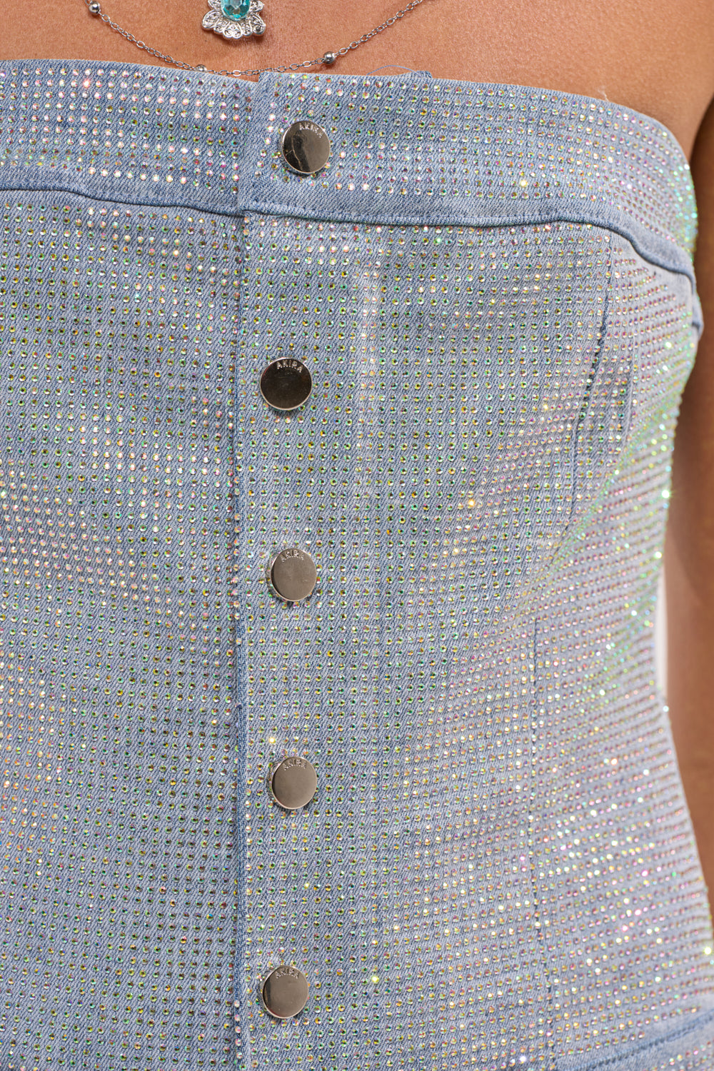 Close-up of the TOO FLY RHINESTONE DENIM JUMPSUIT—a light blue, strapless denim top detailed with metallic buttons and sparkling rhinestones. A turquoise pendant necklace is partially visible at the neckline. 8