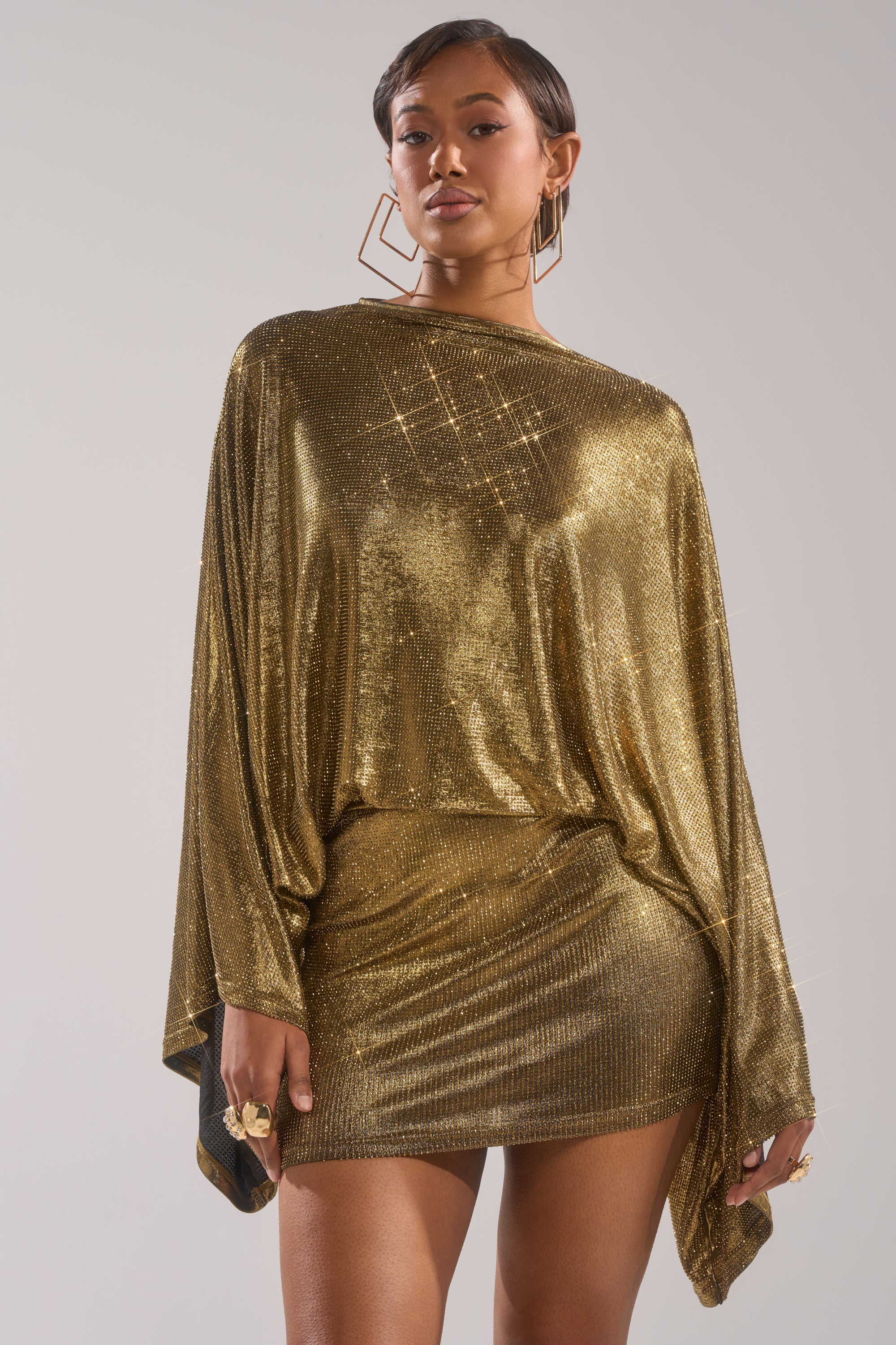 A woman models the GOLDEN HOUR RHINESTONE MINI DRESS, featuring long sleeves, a loose top with a fitted skirt. She accessorizes with large geometric earrings and rings, her short hair styled confidently against a plain backdrop.