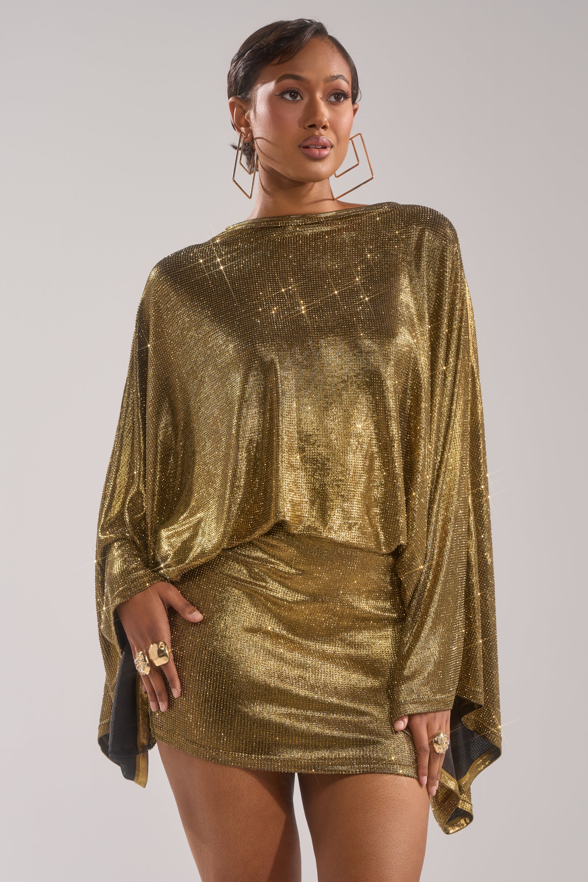 A woman poses confidently in the GOLDEN HOUR RHINESTONE MINI DRESS with long wide sleeves and a short hemline, styled with large square earrings and rings, her hair slicked back against a neutral background.