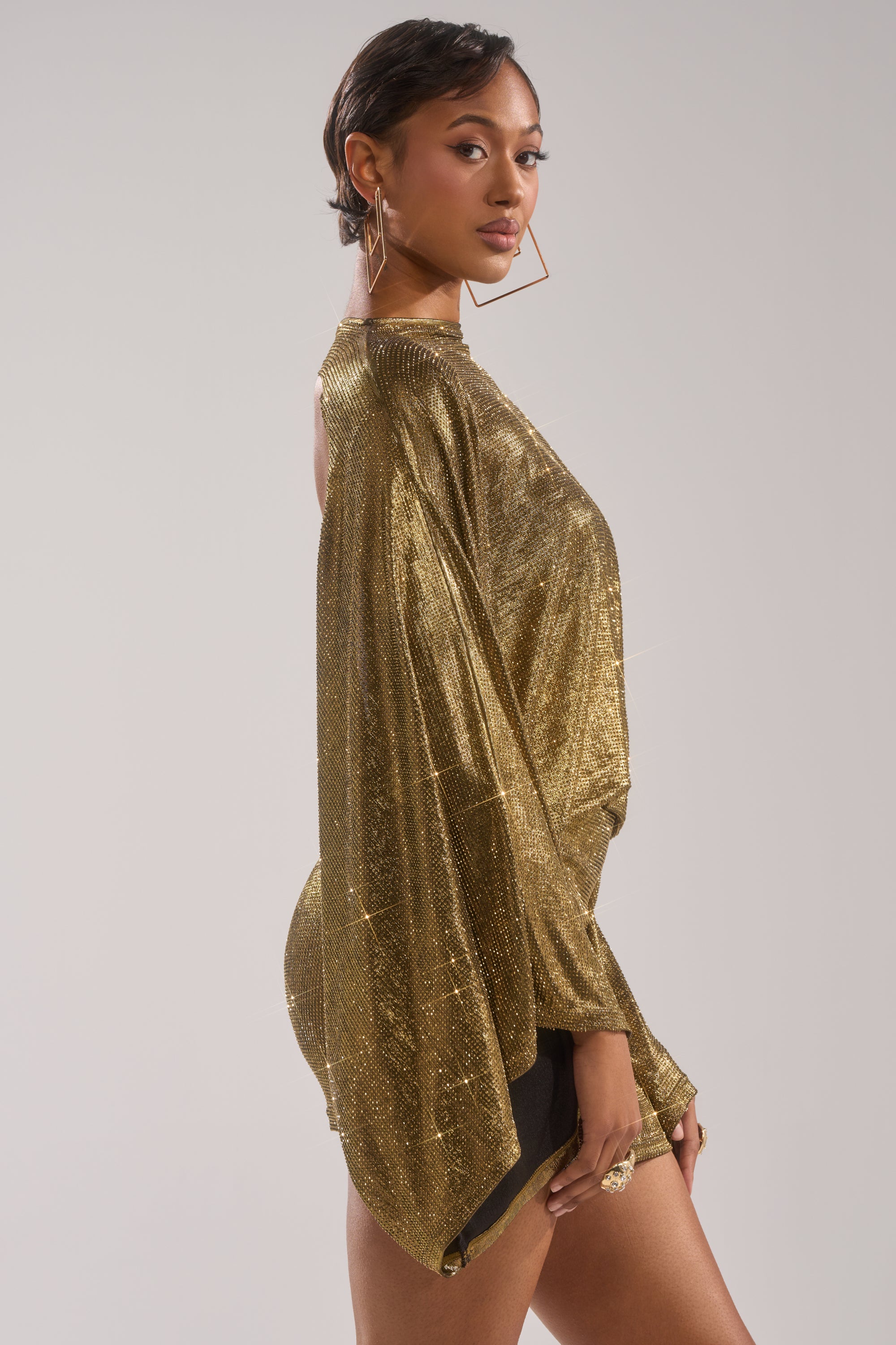 A woman with short hair poses sideways in the GOLDEN HOUR RHINESTONE MINI DRESS with loose, flowing sleeves, styled with geometric gold earrings and a ring, set against a plain light background.