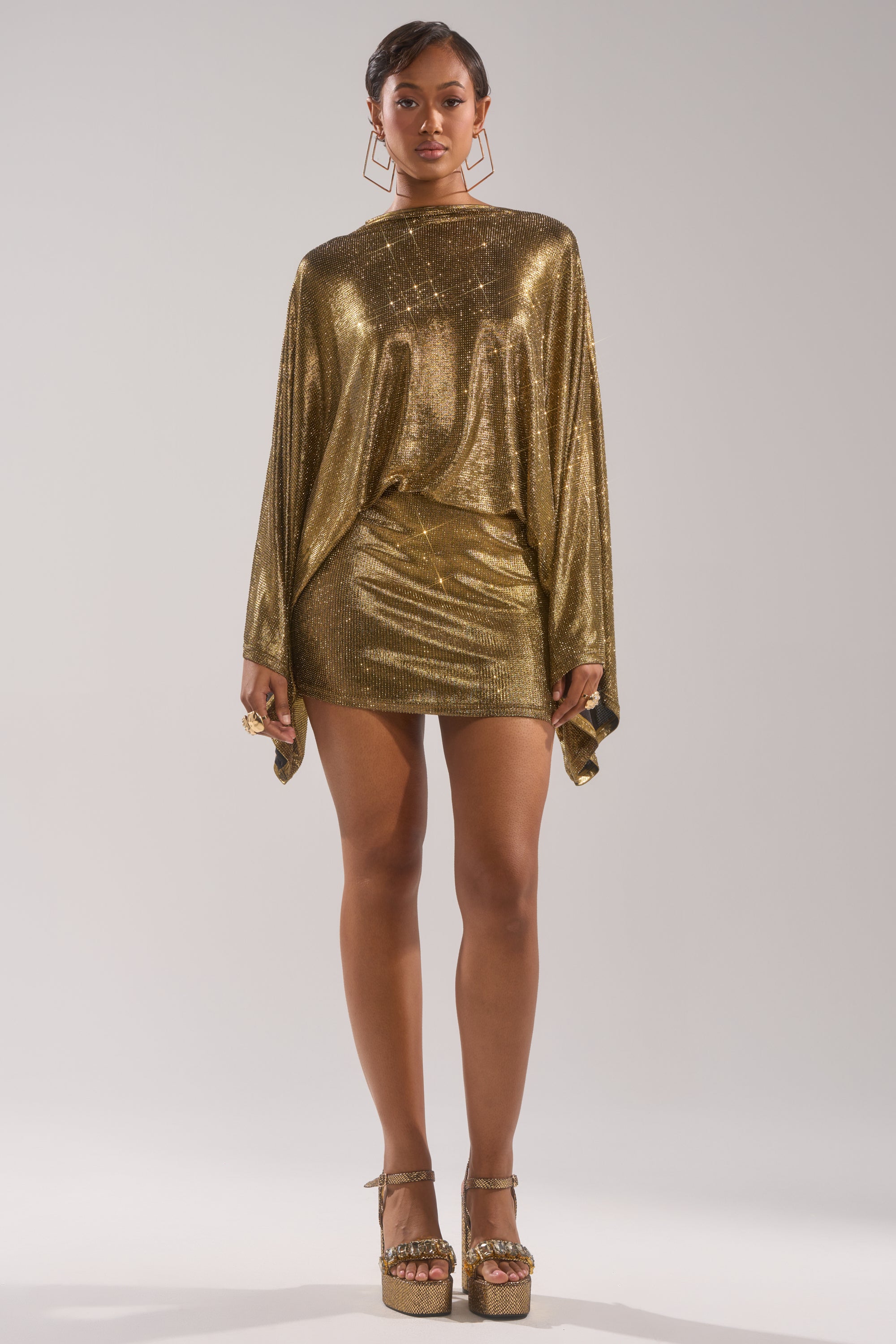 A woman with short hair and gold geometric earrings wears the GOLDEN HOUR RHINESTONE MINI DRESS with draped sleeves and platform heels, standing against a plain light background with a neutral expression.