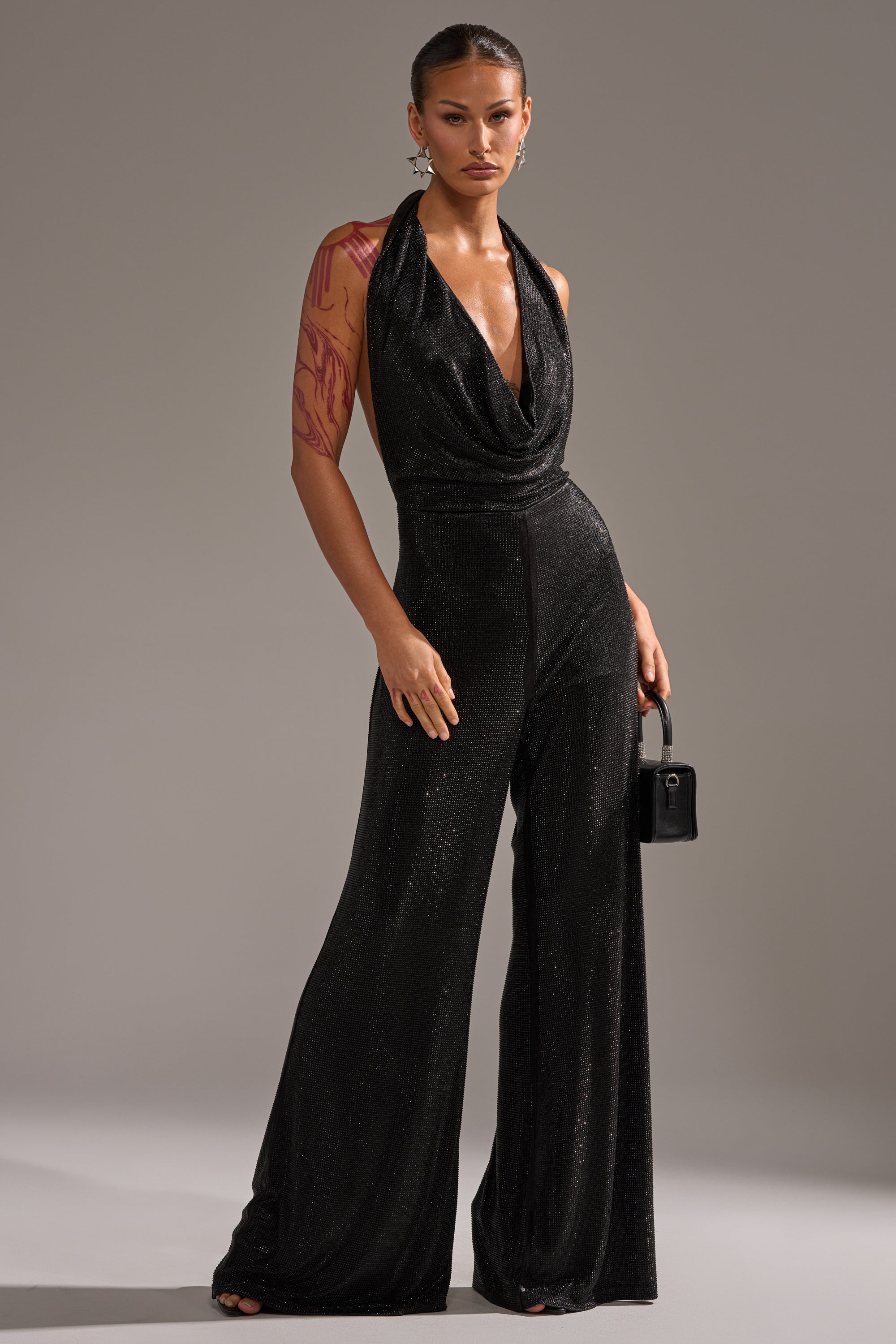 A woman stands confidently in the MAKE YOU DO A DOUBLE TAKE RHINESTONE JUMPSUIT IN BLACK with a draped neckline, holding a black handbag. She has slicked-back hair, bold earrings, and a red tattoo sleeve on her right arm.