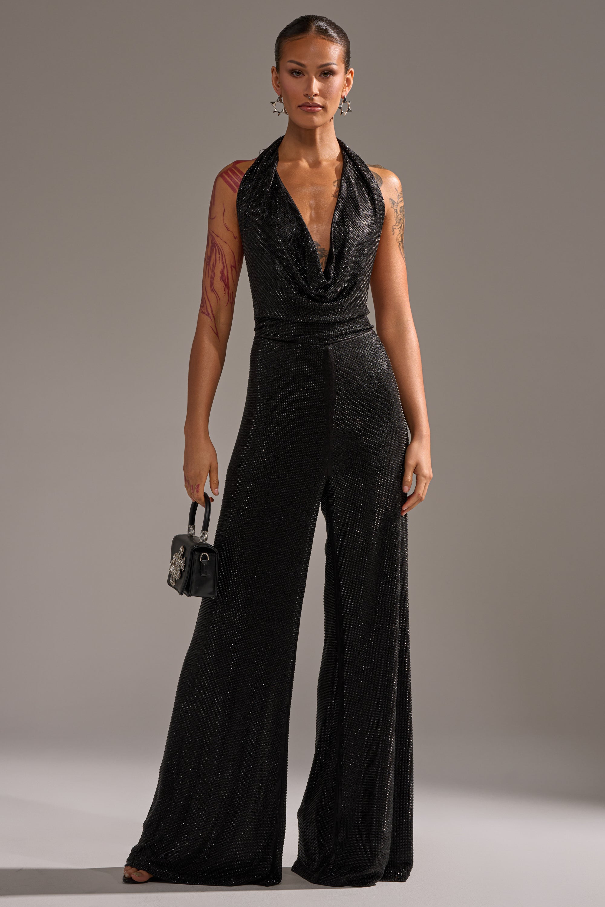 A woman stands confidently in the MAKE YOU DO A DOUBLE TAKE RHINESTONE JUMPSUIT IN BLACK, a sleeveless halter-neck style. She holds a small black handbag, wears hoop earrings, has tattoos on her arm, and her hair is pulled back against a plain gray background.