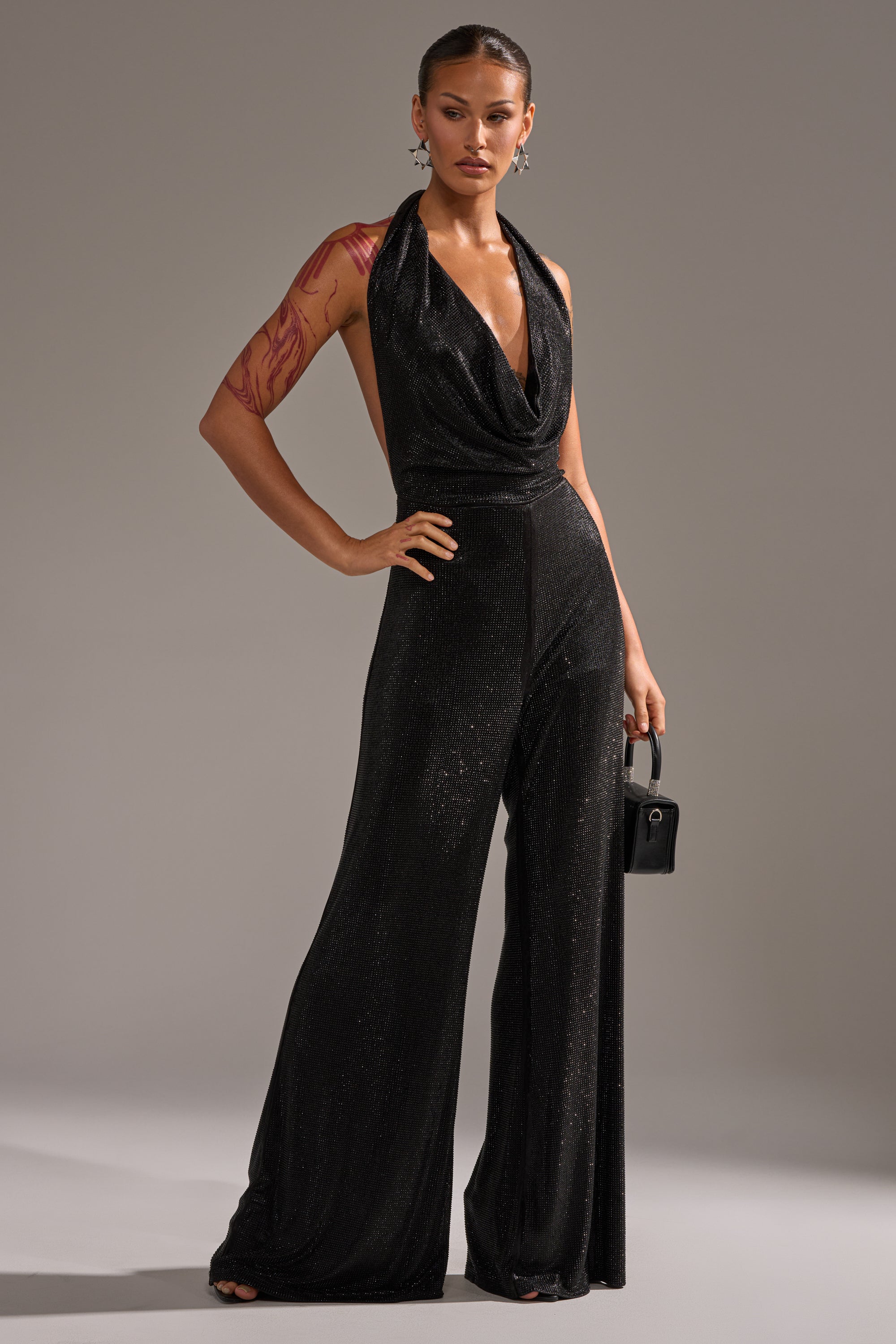 A person stands confidently in the MAKE YOU DO A DOUBLE TAKE RHINESTONE JUMPSUIT IN BLACK, paired with a small black handbag. Their hair is slicked back, they wear statement earrings, and have a red body art design on their left arm against a gray background.