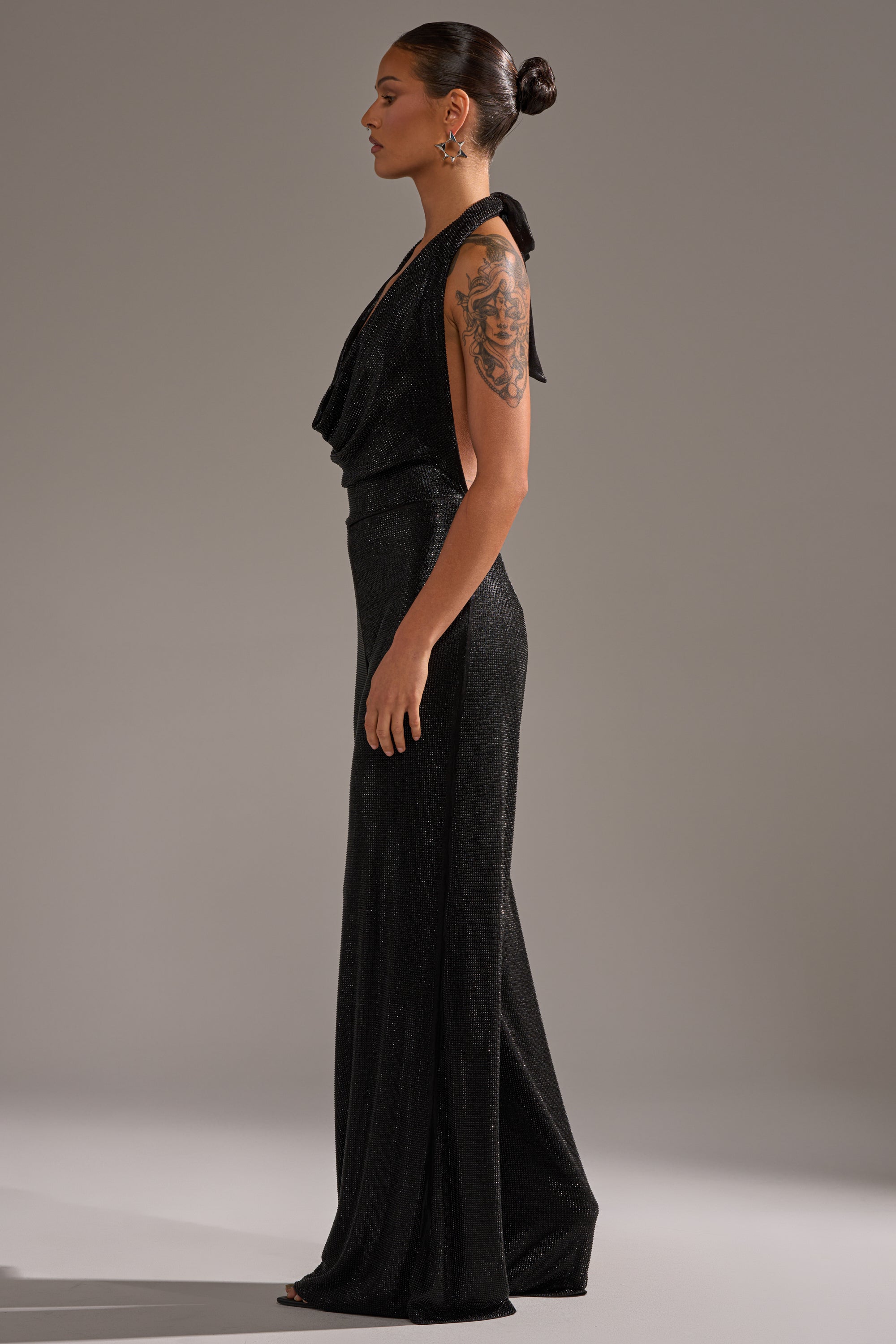 A woman stands in profile wearing the MAKE YOU DO A DOUBLE TAKE RHINESTONE JUMPSUIT IN BLACK. Her hair is in a sleek bun, she has a visible arm tattoo and earrings, and the background is plain gray.