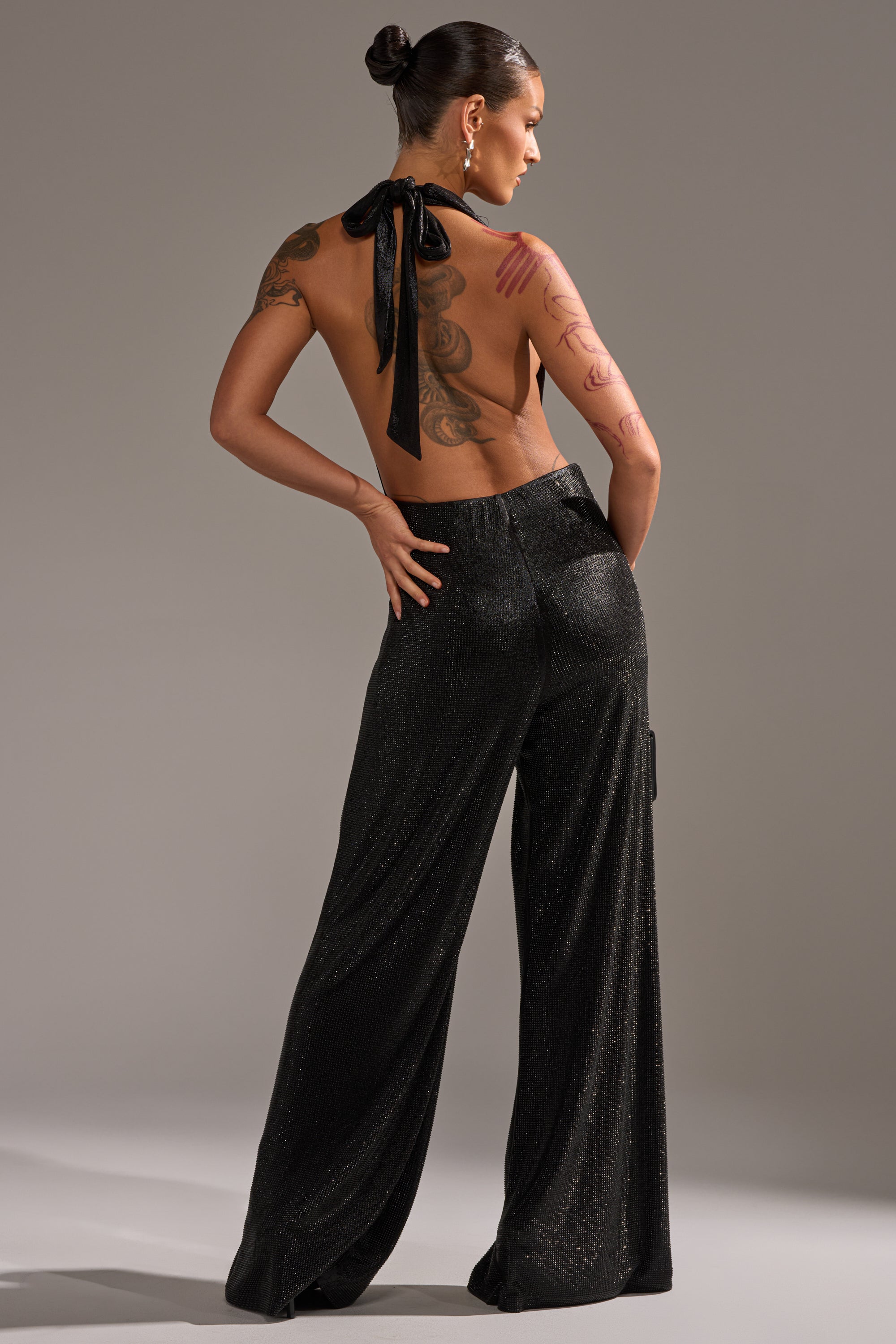 A woman with her hair in a neat bun shows off her tattoos as she stands with her back to the camera, wearing the MAKE YOU DO A DOUBLE TAKE RHINESTONE JUMPSUIT IN BLACK featuring sparkling wide-leg pants and a halter neck that ties, revealing her bare back.