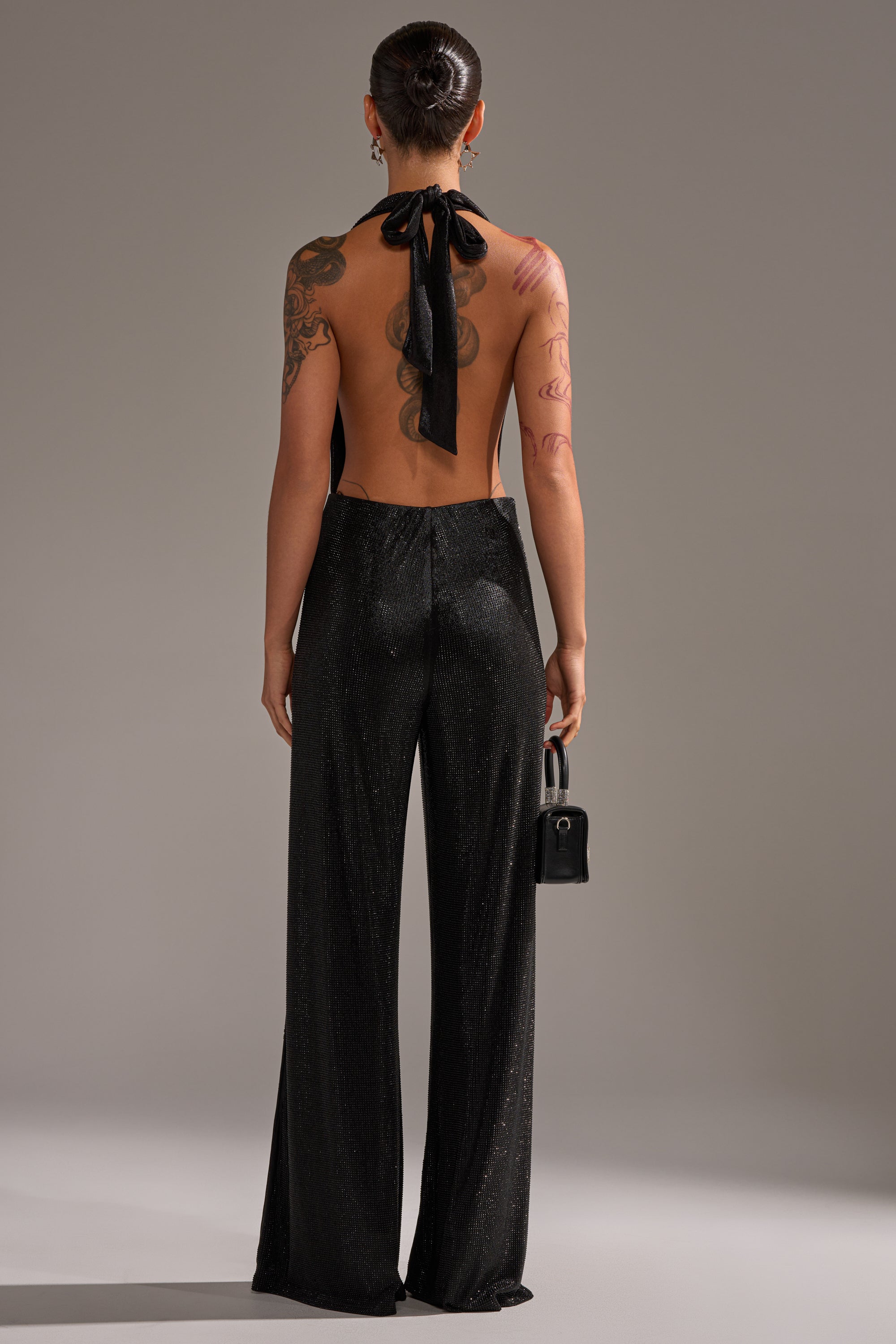 A tattooed person is seen from behind, wearing the MAKE YOU DO A DOUBLE TAKE RHINESTONE JUMPSUIT IN BLACK with a low back. They hold a small black handbag and their hair is styled in a sleek bun.