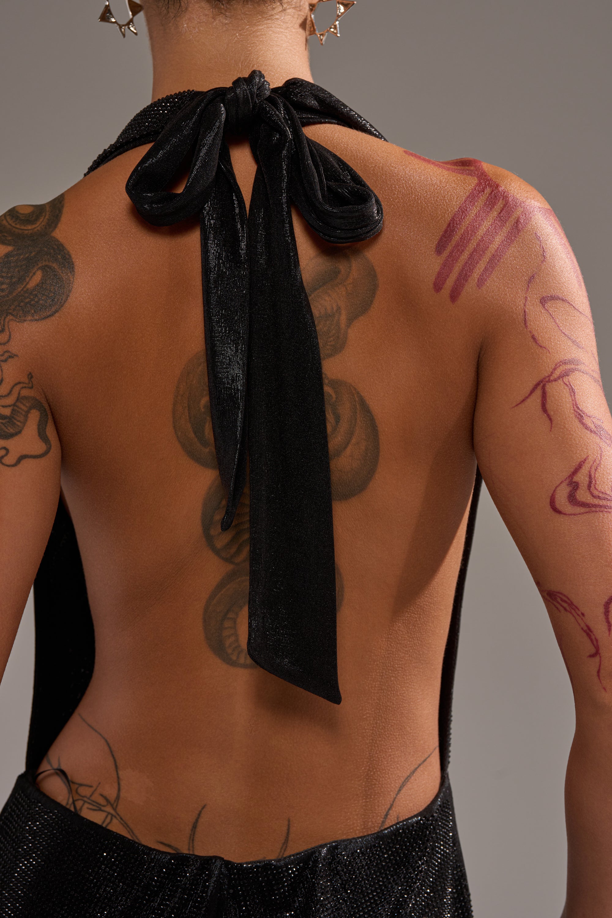 A person with tattoos and red body paint on their back and arm wears the MAKE YOU DO A DOUBLE TAKE RHINESTONE JUMPSUIT IN BLACK, a sparkly, backless outfit tied with a bow at the neck. The background is plain and neutral.