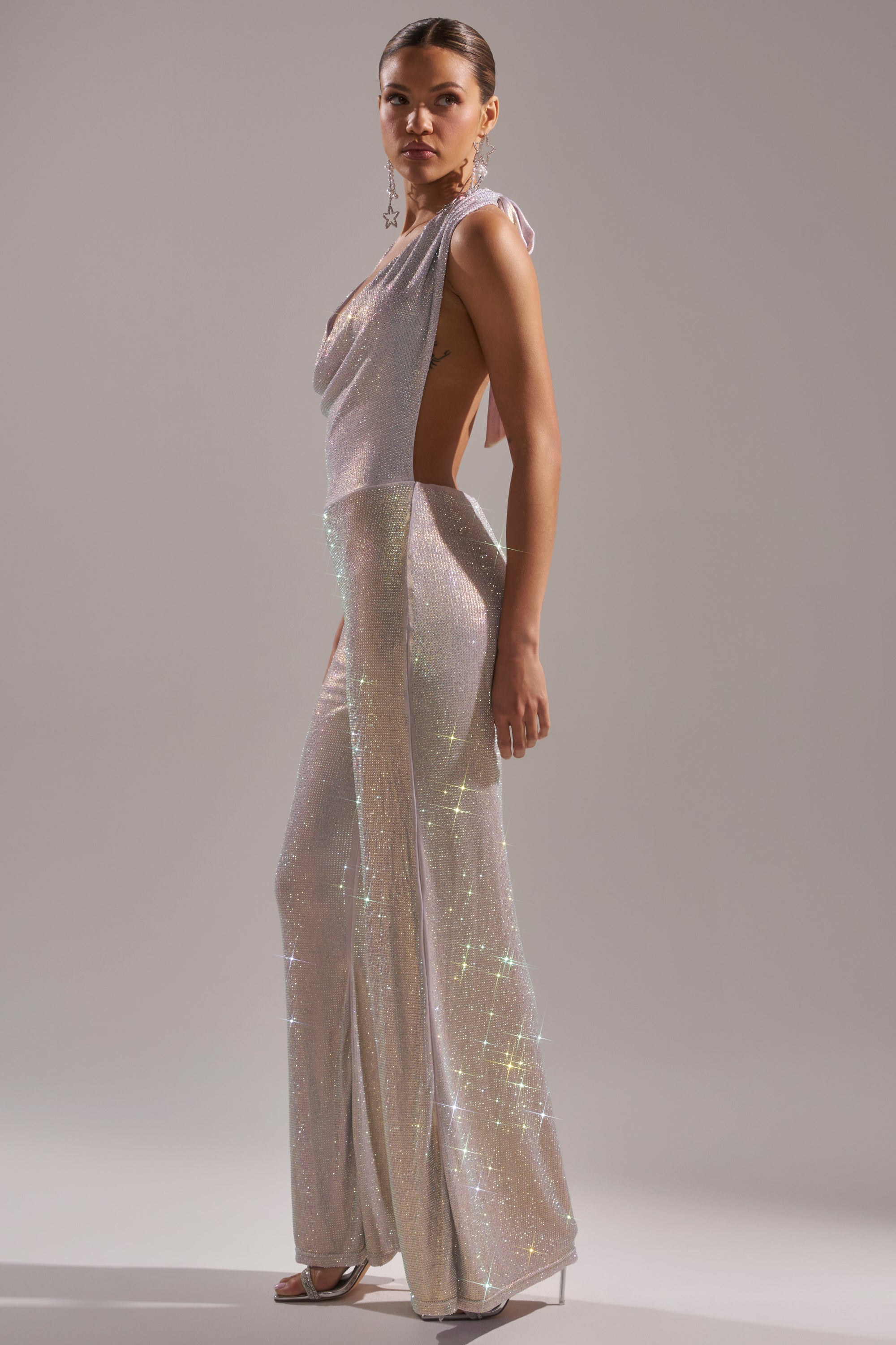 A woman poses in profile against a plain gray background wearing the MAKE YOU DO A DOUBLE TAKE Rhinestone Jumpsuit in Champagne, featuring a shimmery, sleeveless, backless design with a draped neckline and metallic heels.