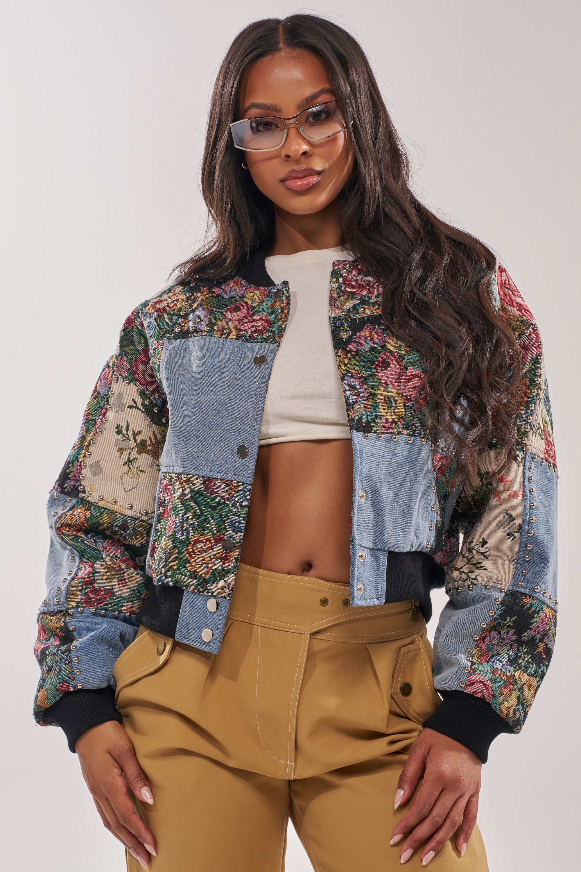 A woman confidently poses with her hands in her pockets against a plain background, wearing clear glasses and the SWEET TALKER PATCHWORK BOMBER over a white crop top with high-waisted tan pants. 0