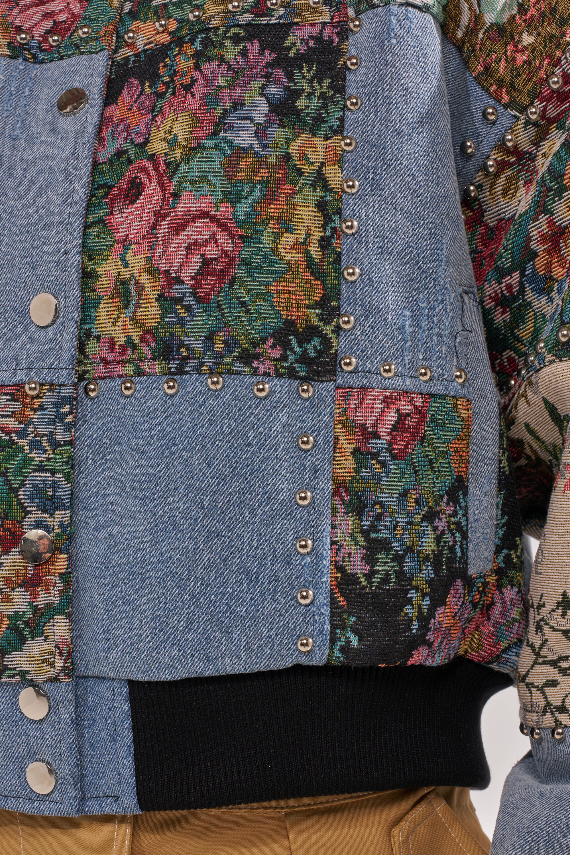 Close-up of the SWEET TALKER PATCHWORK BOMBER, featuring denim and floral tapestry panels with metal studs on the seams. A black ribbed waistband lines the bottom edge, paired with tan pants for a street-chic style. 9