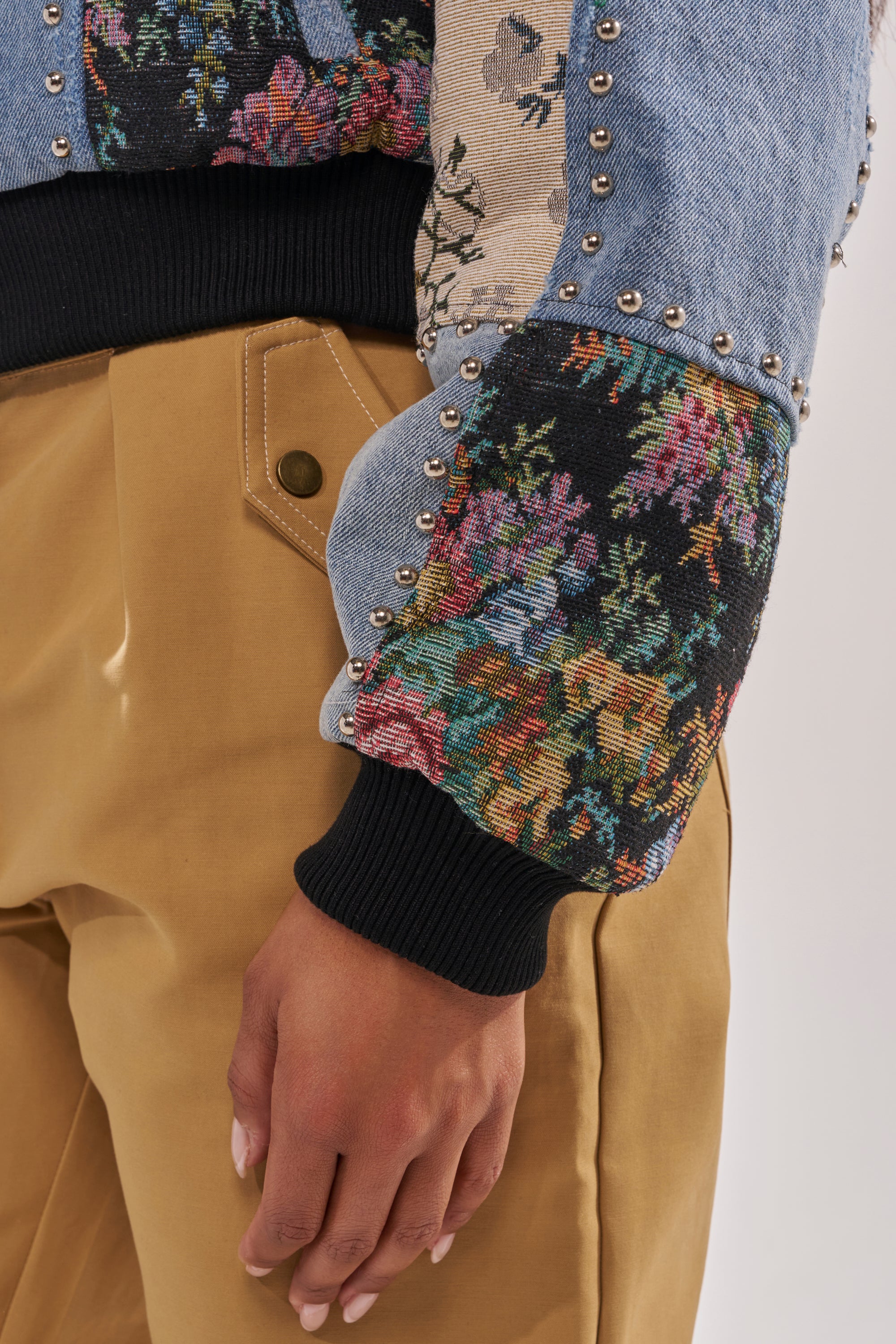A close-up of a person wearing tan pants and the SWEET TALKER PATCHWORK BOMBER, a street-chic jacket with denim, floral tapestry fabric, and metal studs on the sleeve. Their hand rests by their side. 10