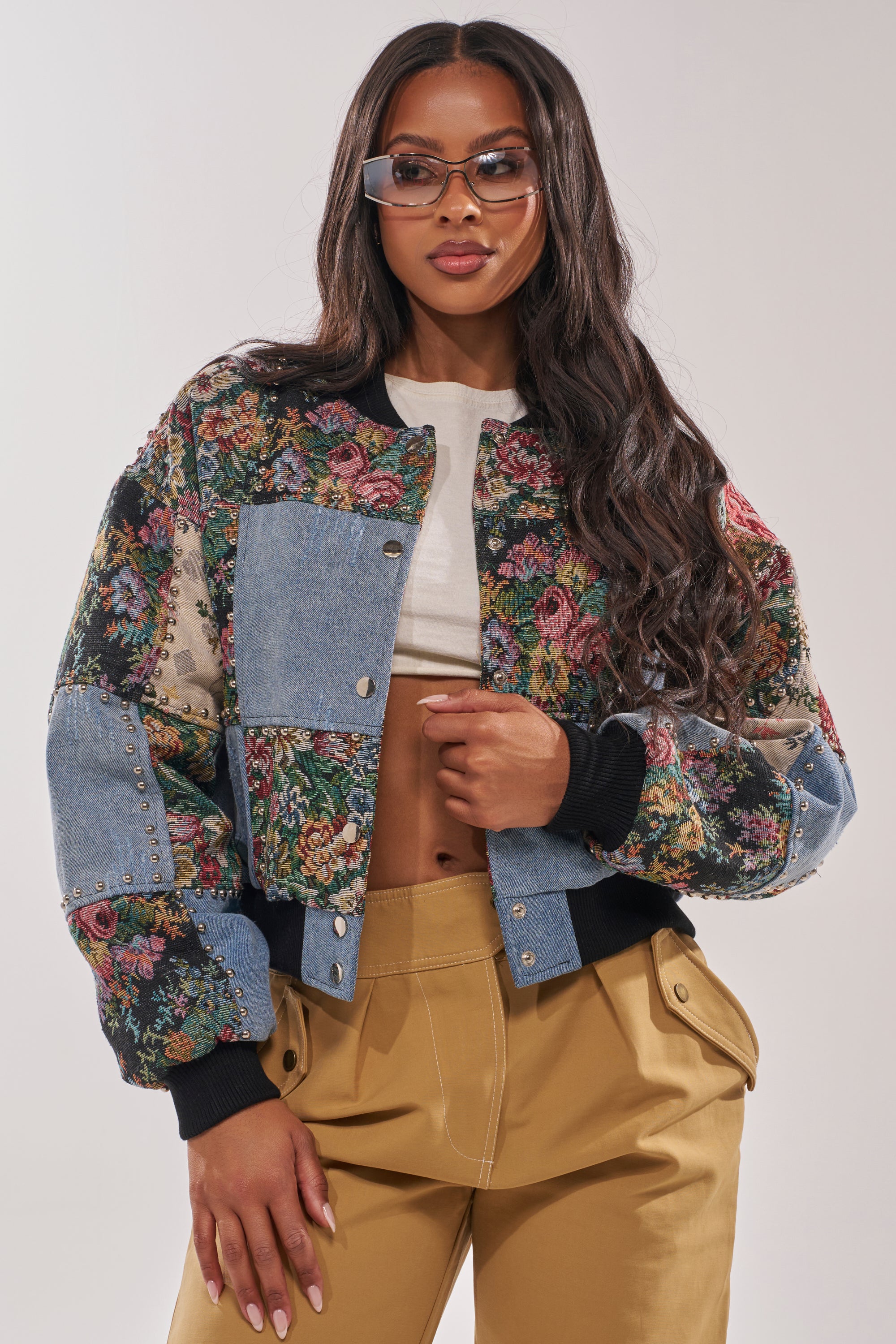 A woman poses confidently in the SWEET TALKER PATCHWORK BOMBER, featuring a tapestry floral design with denim patchwork, paired with tan pants, a white cropped top, and clear sunglasses against a plain background. 2