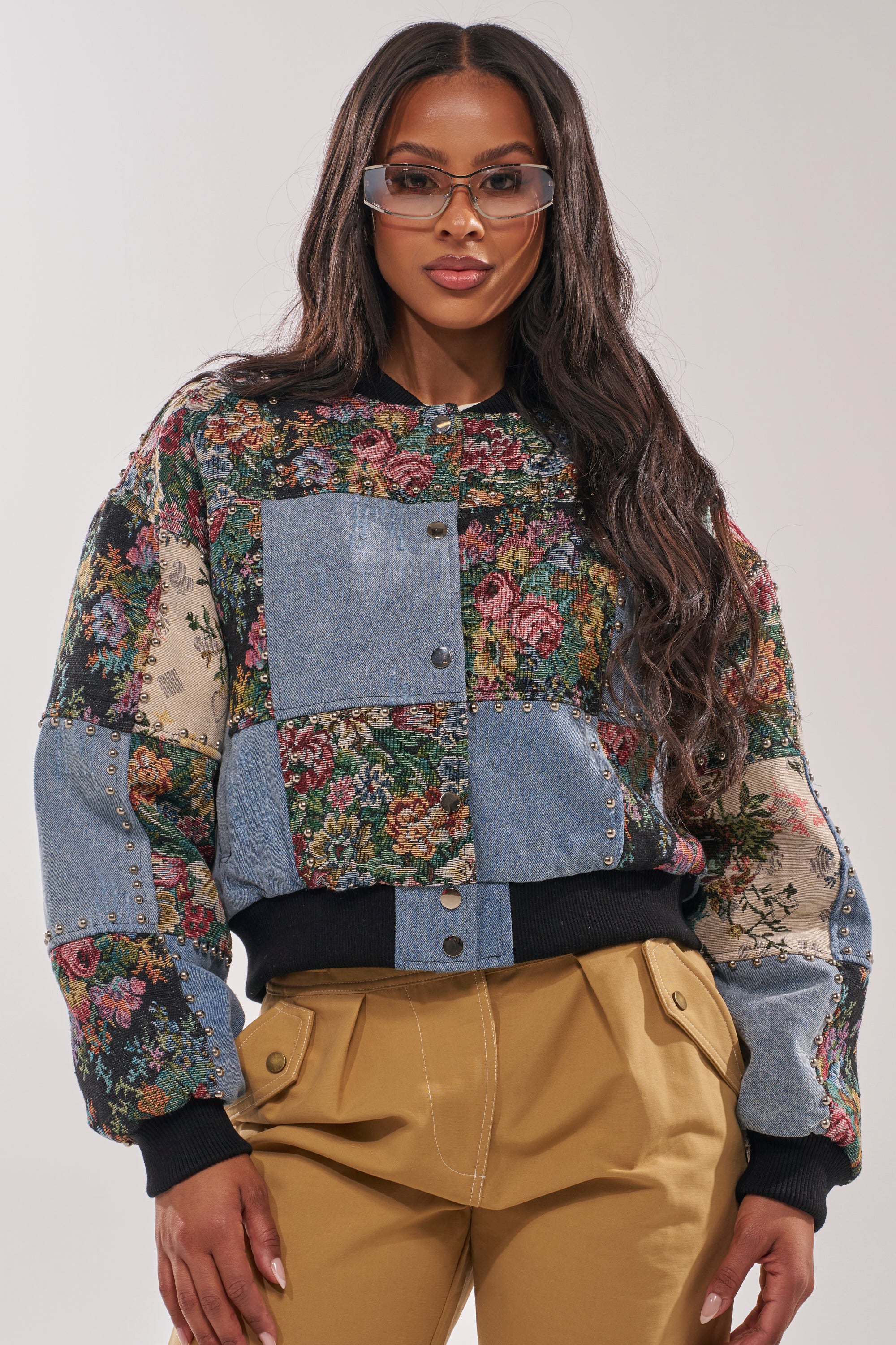 A woman in sunglasses and tan high-waisted pants models the SWEET TALKER PATCHWORK BOMBER, featuring tapestry florals and denim patchwork, against a plain light background. 3