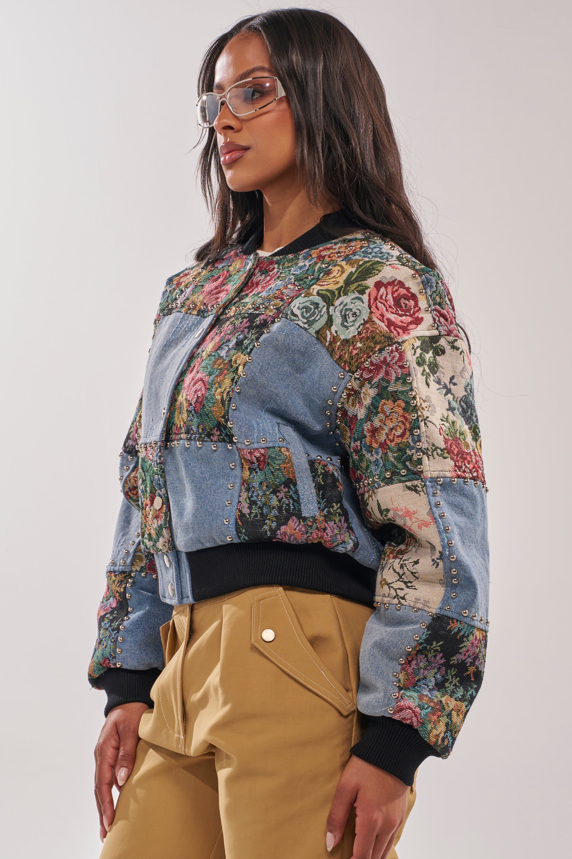 A woman with long dark hair and clear glasses wears the SWEET TALKER PATCHWORK BOMBER—a tapestry floral jacket with stud details—and tan pants, standing against a plain background. 4