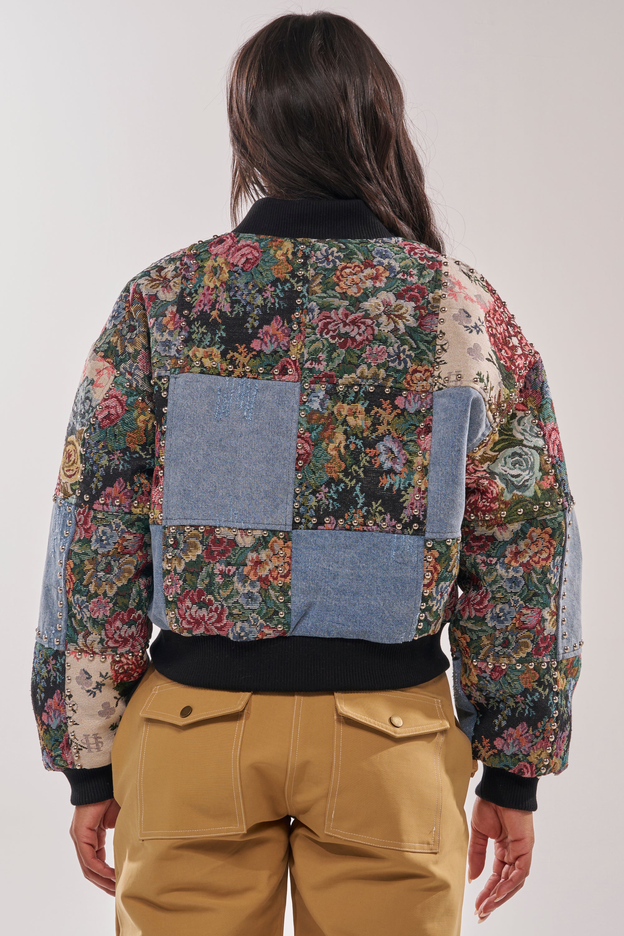 A person with long dark hair is seen from behind wearing the SWEET TALKER PATCHWORK BOMBER—a vibrant tapestry jacket mixing colorful florals and classic blue denim patchwork—paired with tan pants for a street-chic look. 5