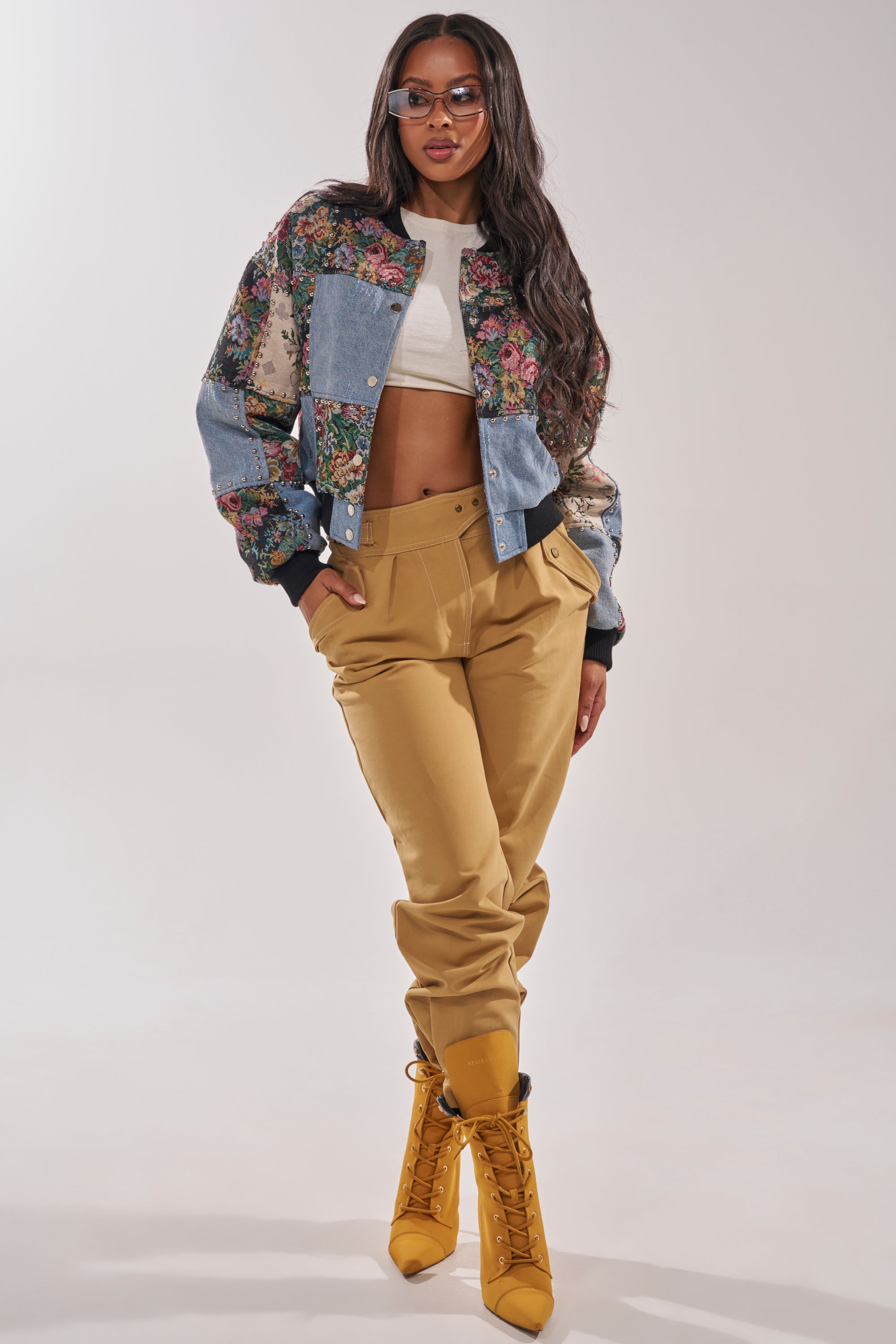 A woman stands confidently against a light background, wearing sunglasses and the SWEET TALKER PATCHWORK BOMBER over a white crop top, paired with tan high-waisted pants and mustard yellow lace-up boots. 6