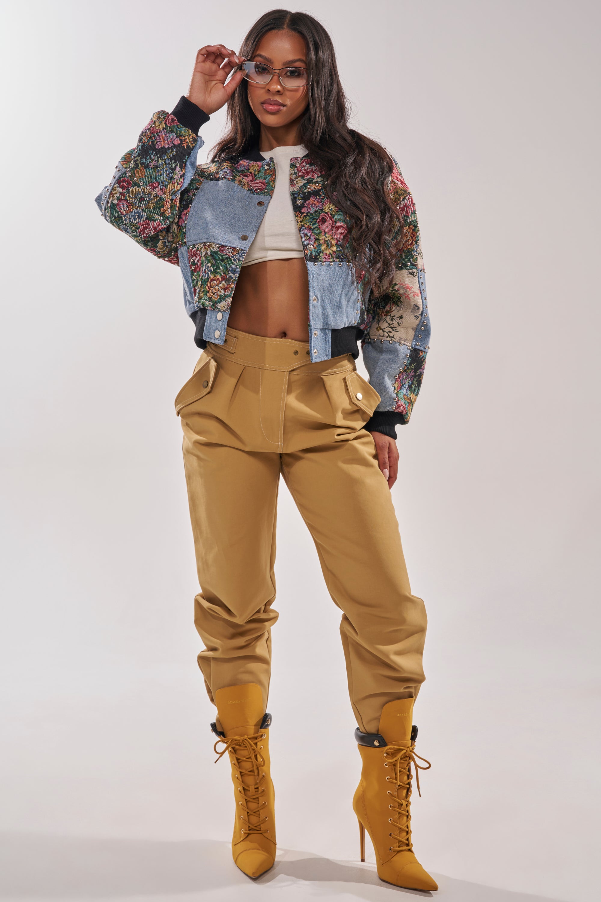 A woman poses confidently in the SWEET TALKER PATCHWORK BOMBER, paired with a white crop top, high-waisted tan pants, and mustard yellow lace-up boots. She wears sunglasses and has long wavy hair against a plain background. 7