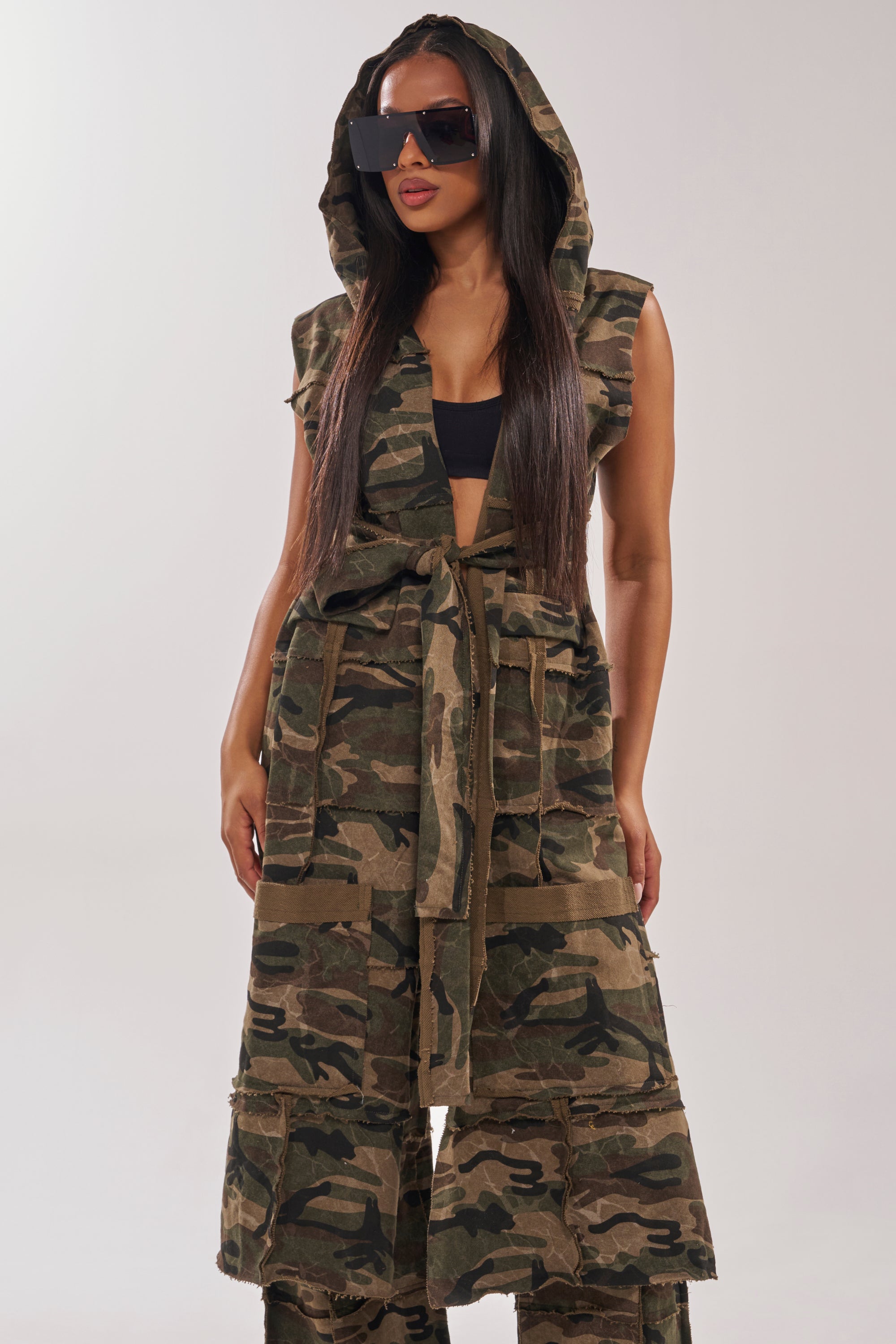 A woman wears the CAN'T SEE ME HOODED LONGLINE CAMO SWEATSHIRT with a tie belt, styled over a black crop top and matching pants. She has long straight hair and large black sunglasses. 0