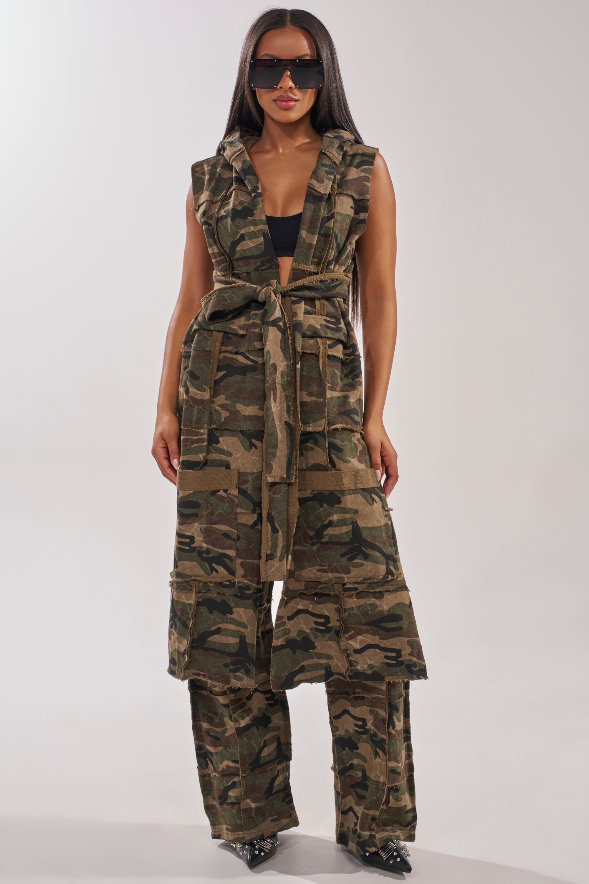 A woman stands confidently against a light background wearing the CAN'T SEE ME HOODED LONGLINE CAMO SWEATSHIRT, styled with black shoes, sunglasses, and bold accessories. 1