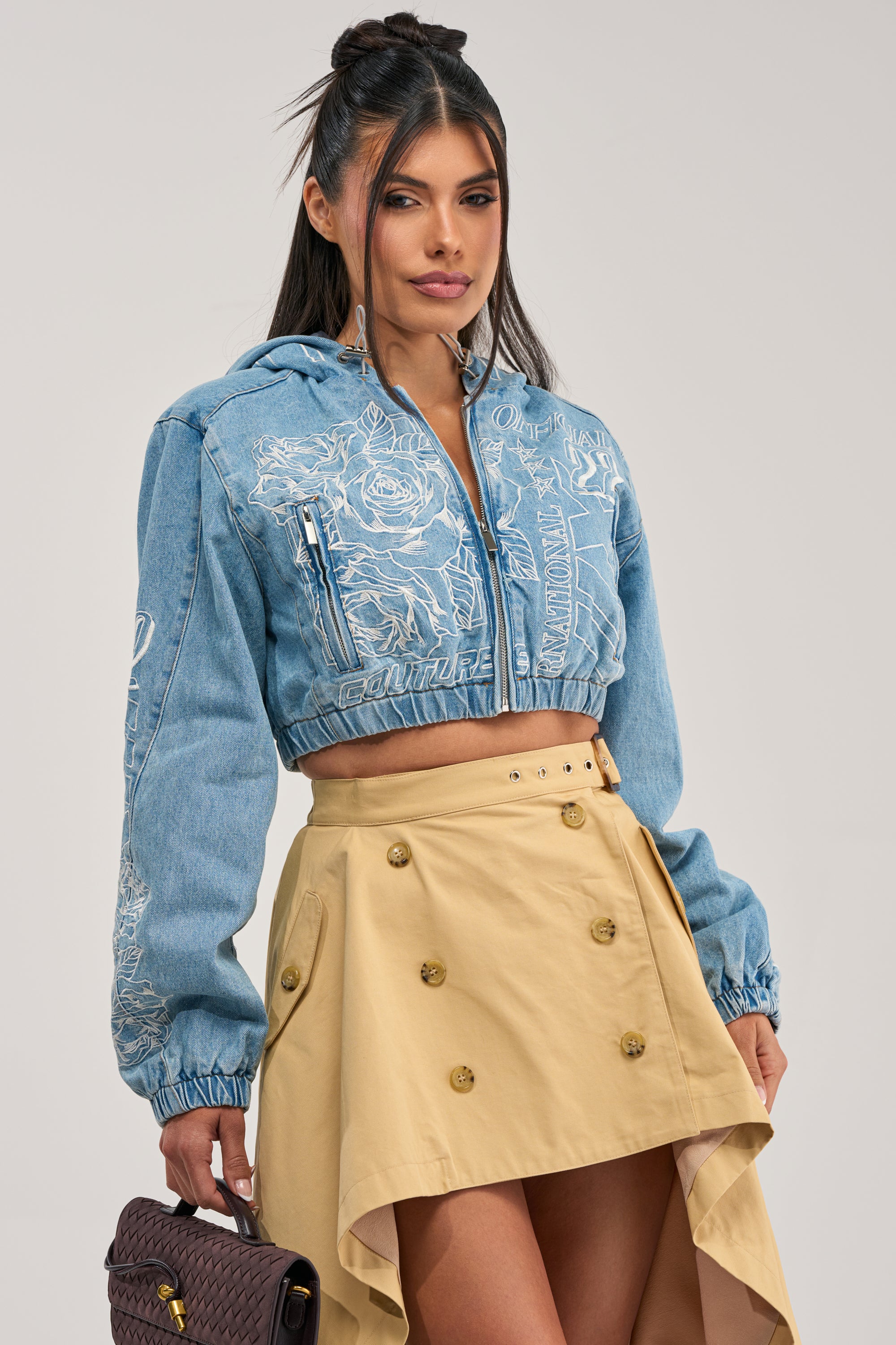 A woman stands against a plain white background, effortlessly chic in the I SPY DENIM BOMBER cropped embroidered jacket and a beige asymmetrical buttoned skirt, holding a brown woven handbag to complete her streetwear look.