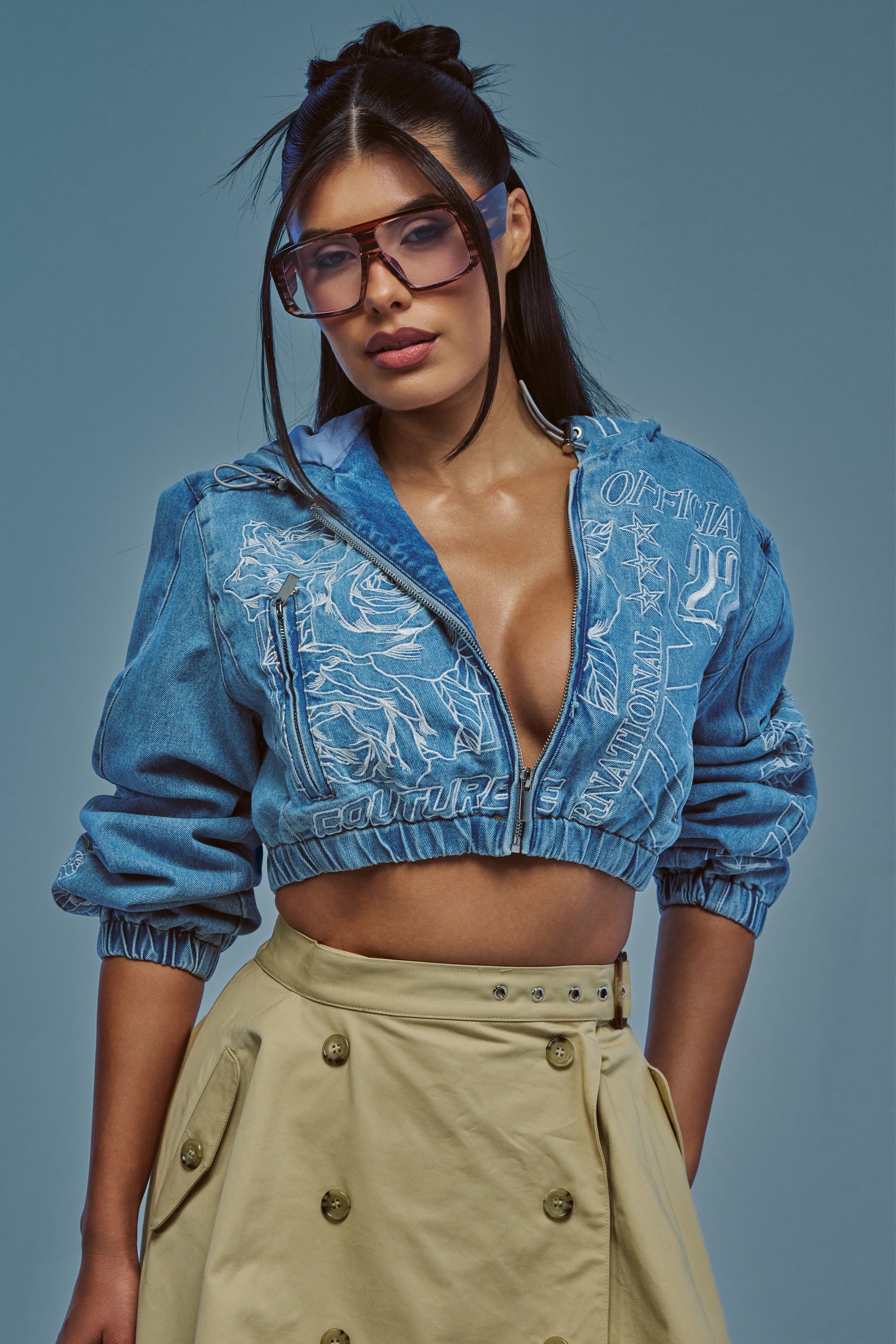Wearing oversized sunglasses and the I SPY DENIM BOMBER—a blue cropped denim jacket with eye-catching embroidery—paired with a beige high-waisted skirt, a woman poses against a plain blue background.