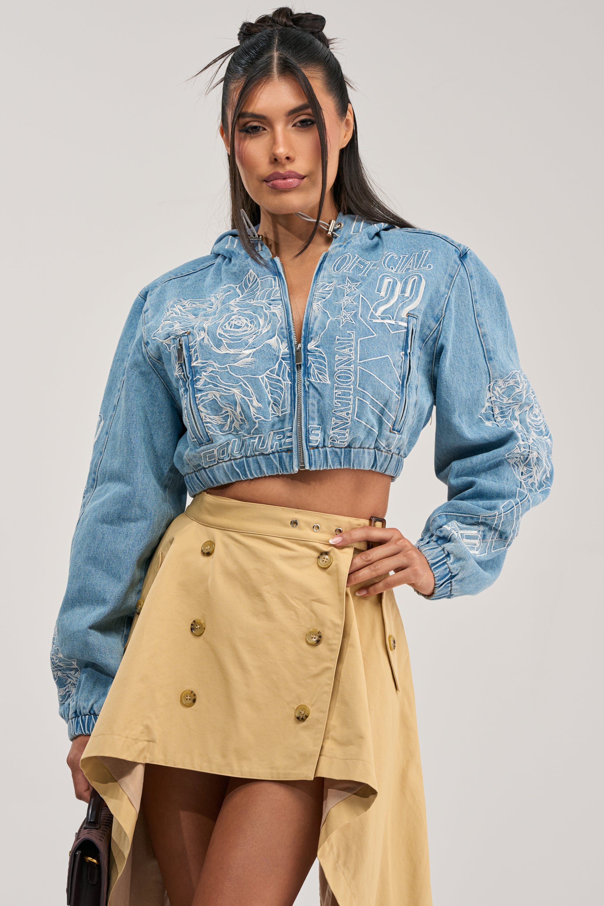 Model wearing the I SPY DENIM BOMBER, a blue cropped jacket featuring white embroidery, styled with a tan asymmetrical buttoned skirt and posing with one hand on her hip against a plain light background for a modern streetwear look.