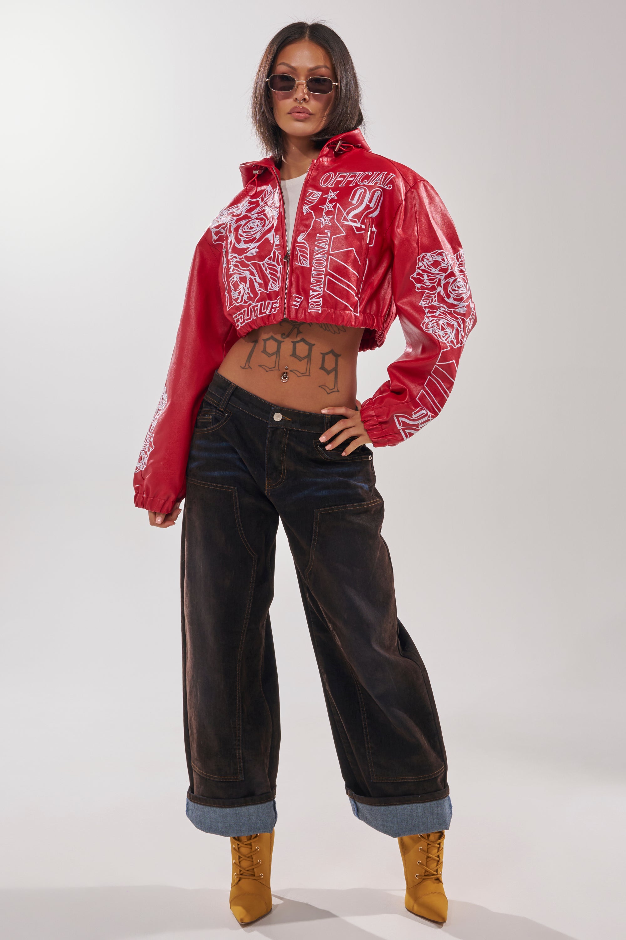A woman stands confidently in the PAINT THE TOWN RED EMBROIDERED BOMBER, loose dark cuffed jeans, yellow boots, sunglasses, and a "1999" tattoo on her stomach. 9
