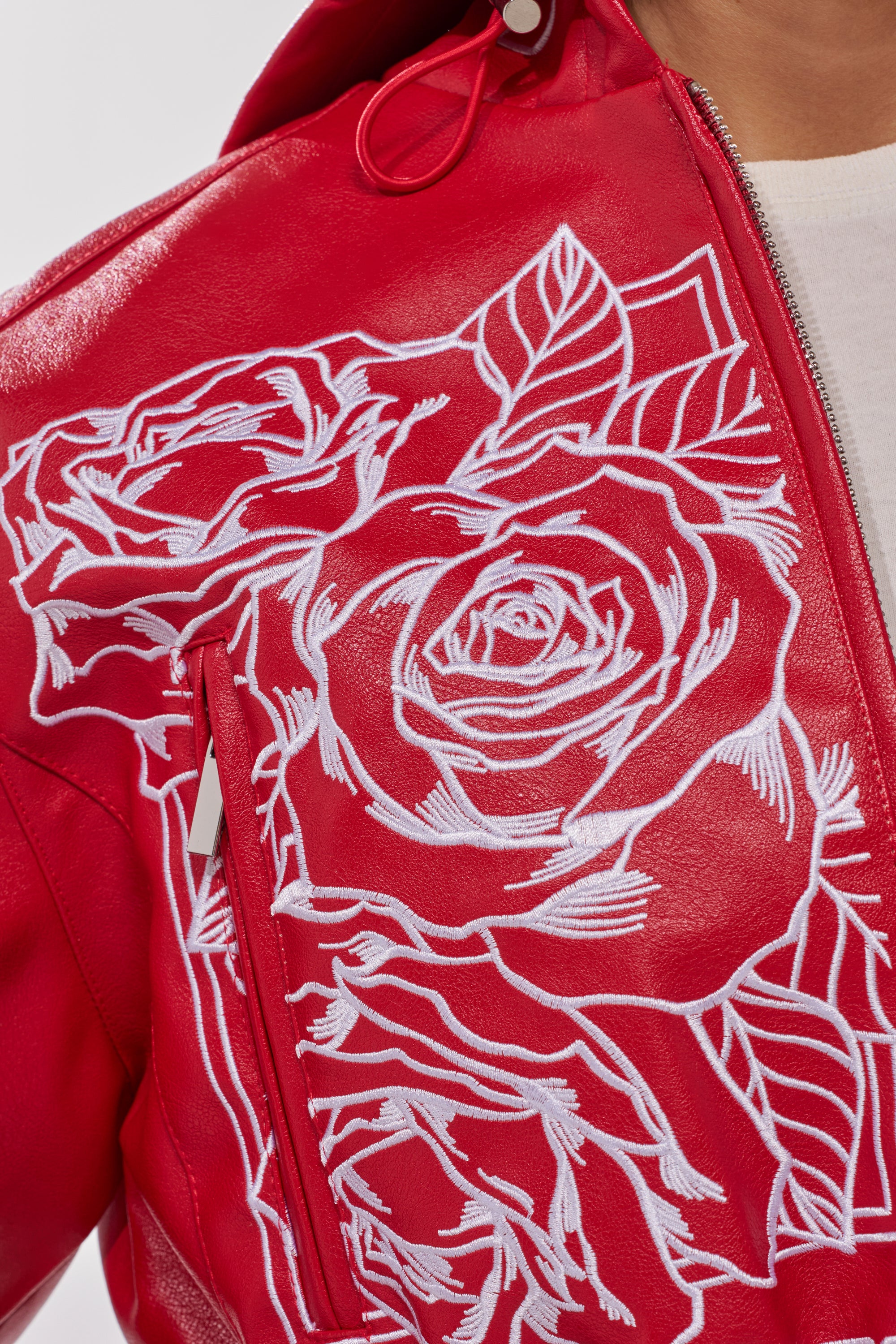A close-up of the PAINT THE TOWN RED EMBROIDERED BOMBER, a red faux leather jacket with bold white rose embroidery, shown partially unzipped to reveal a hint of a white shirt underneath. 10