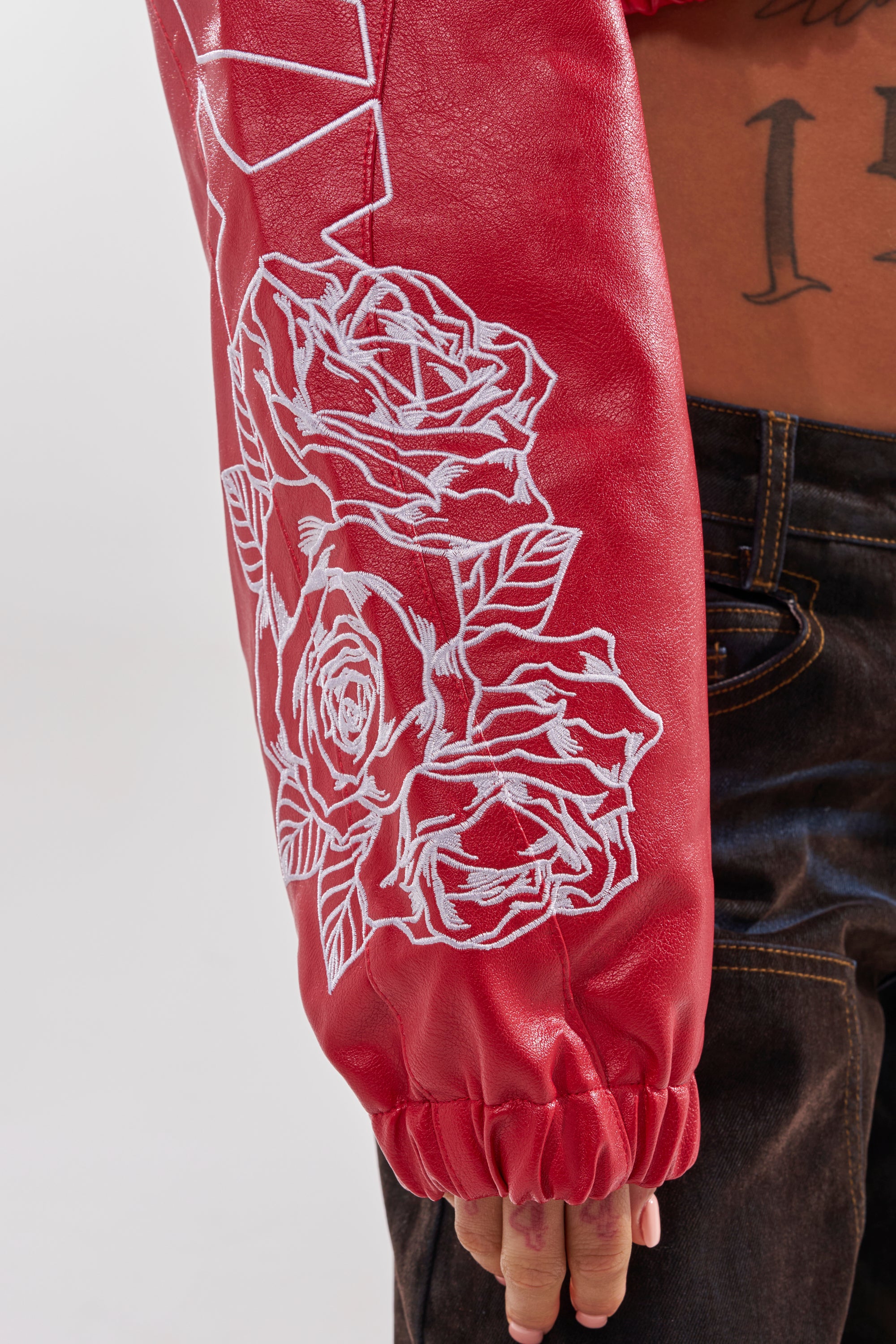 A person exudes a cool streetwear vibe in the PAINT THE TOWN RED EMBROIDERED BOMBER, featuring red faux leather with white rose embroidery on the sleeve, paired with dark jeans and a relaxed pose. 11
