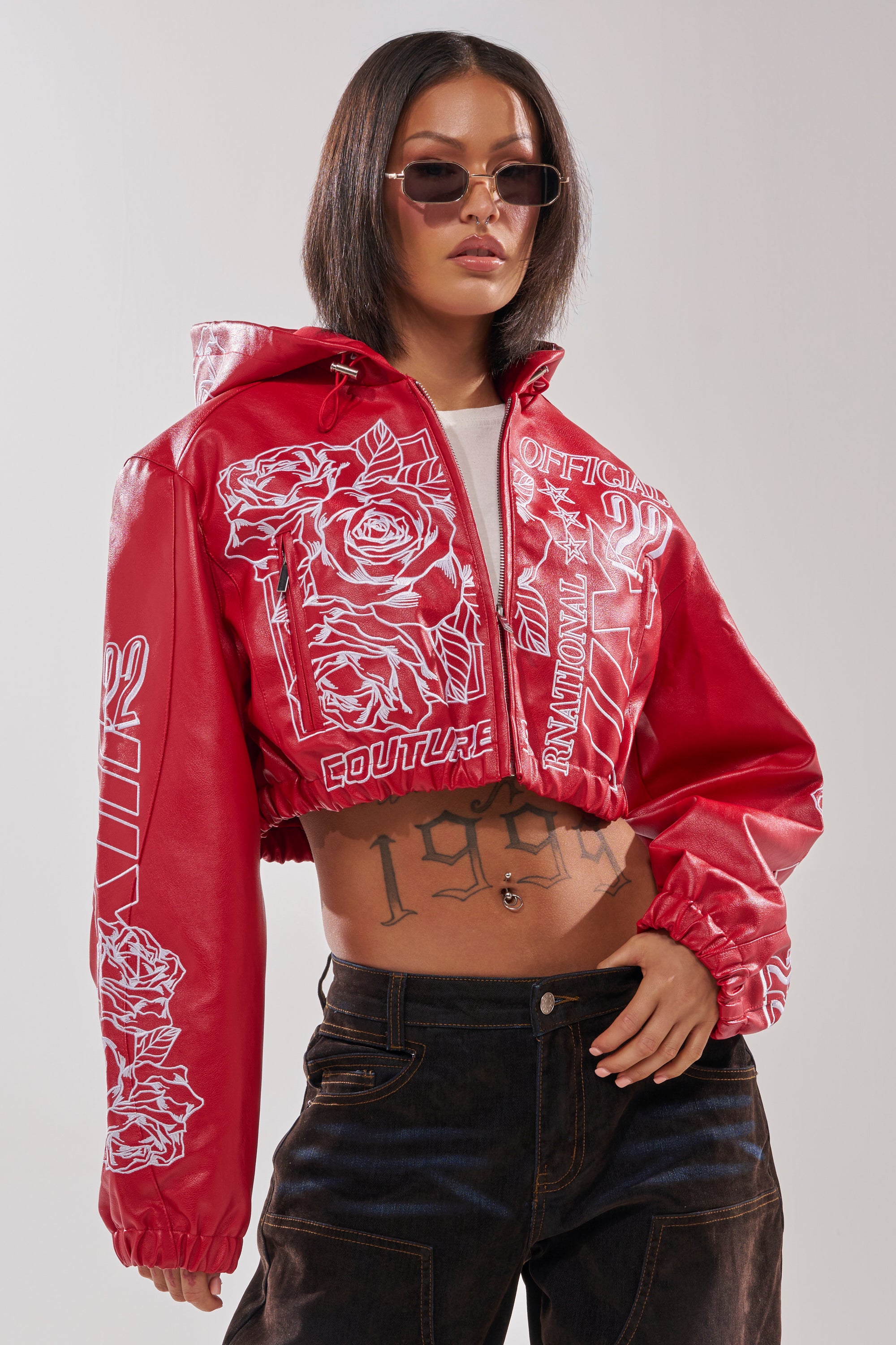 A woman poses confidently in the PAINT THE TOWN RED EMBROIDERED BOMBER, featuring white floral designs, black jeans, sunglasses, and a torso tattoo—delivering a bold modern streetwear look. 1