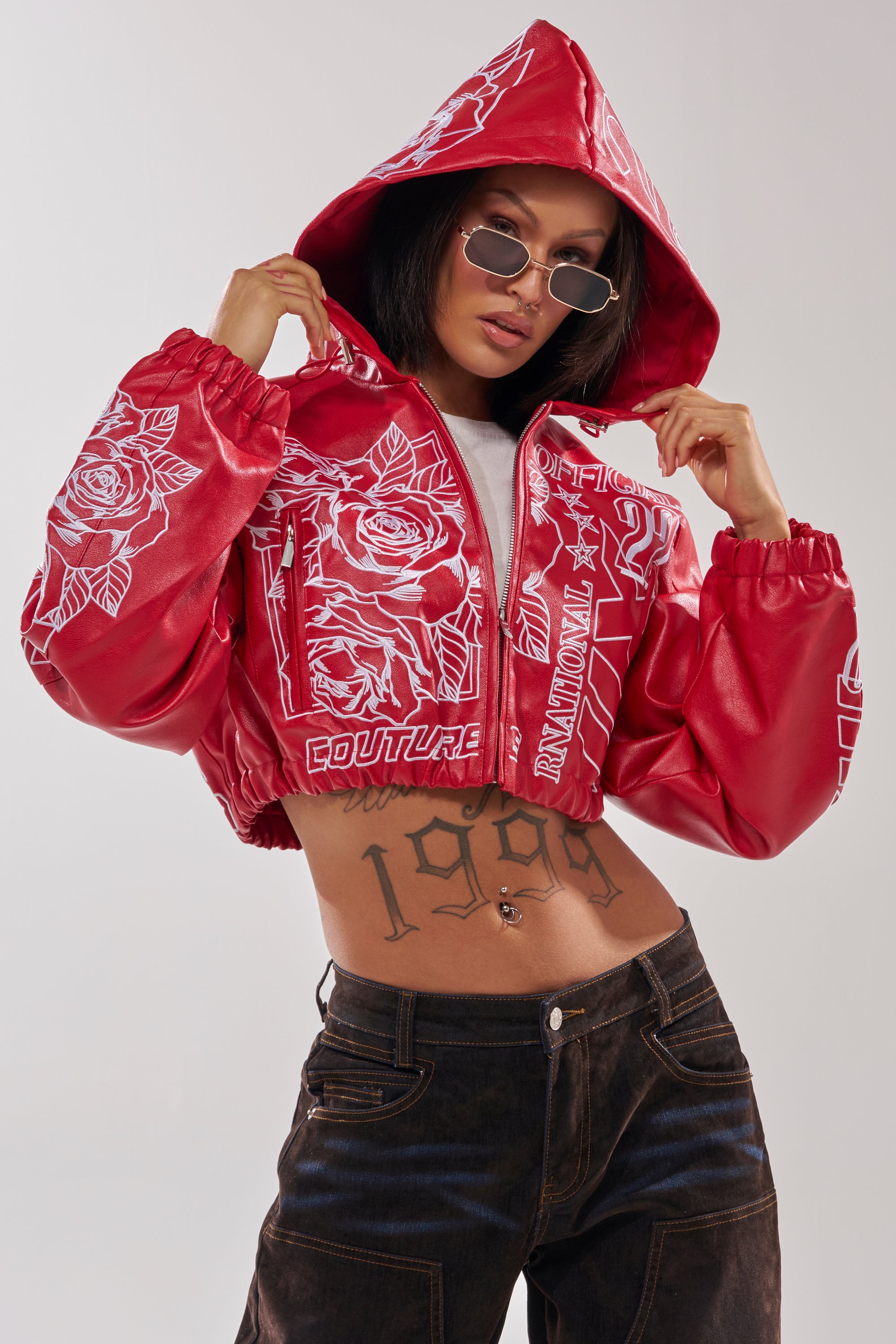 A person in the PAINT THE TOWN RED EMBROIDERED BOMBER, styled as streetwear with white graphics, dark sunglasses, and dark jeans, confidently poses against a plain background, showing a "1989" tattoo across their stomach. 4