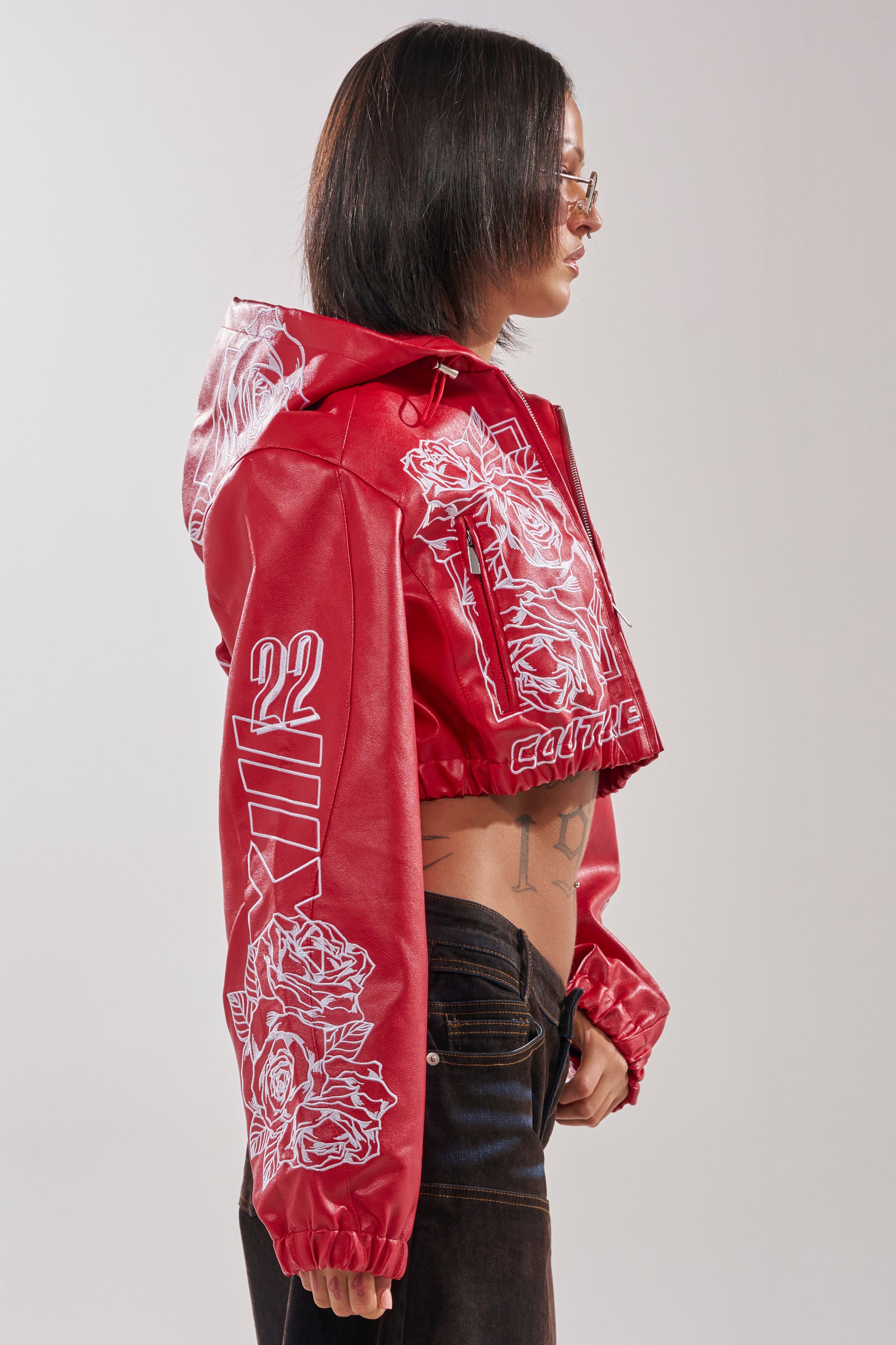 A person with a bob haircut and brown sunglasses wears the PAINT THE TOWN RED EMBROIDERED BOMBER, featuring white rose designs and “22” on the sleeve, paired with black pants, standing in profile against a light background. 5