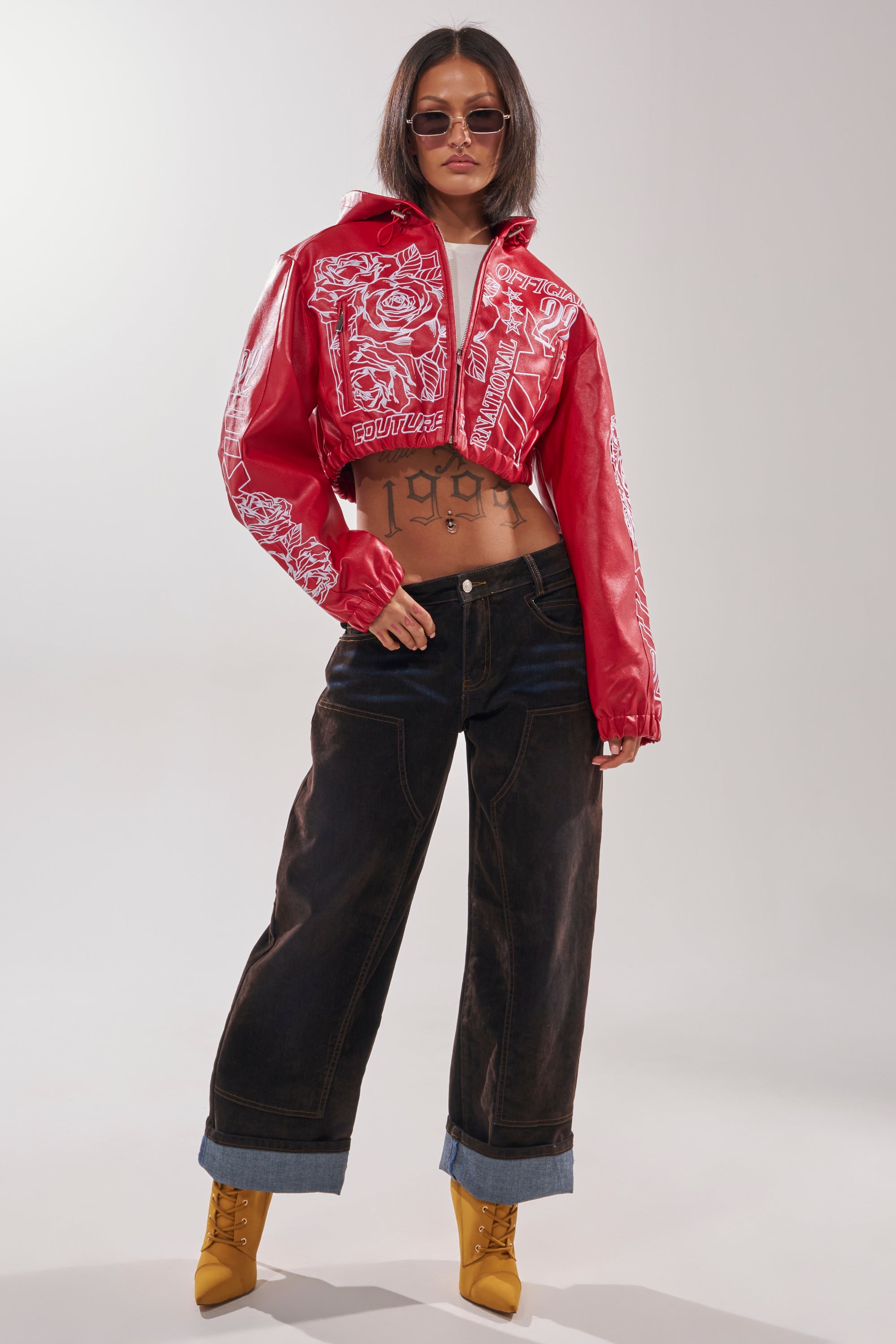 A woman in sunglasses rocks the PAINT THE TOWN RED EMBROIDERED BOMBER with white designs, baggy dark jeans, and yellow heeled boots. Her exposed midriff reveals a “1989” tattoo across her stomach. 7