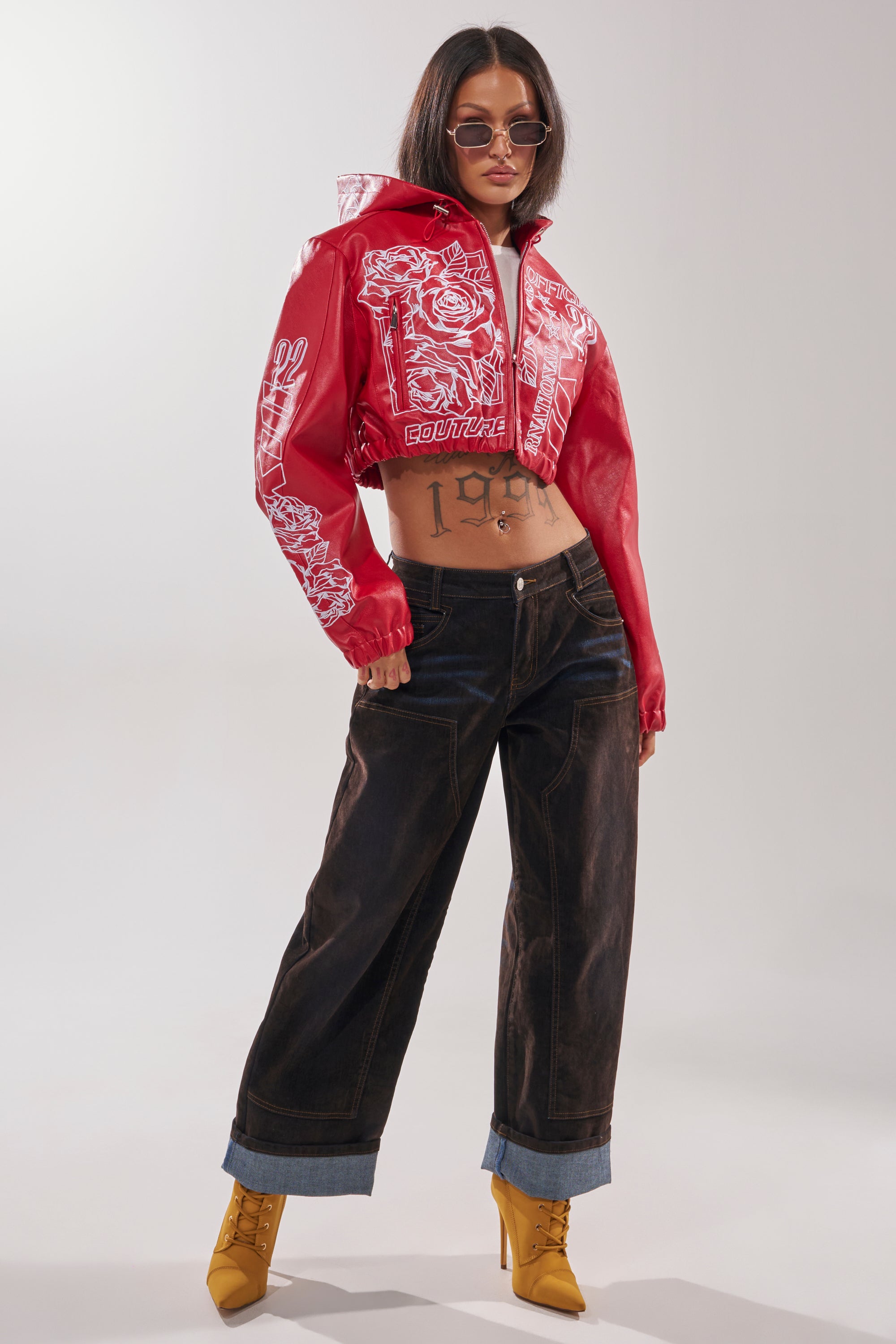 Wearing the PAINT THE TOWN RED EMBROIDERED BOMBER, loose dark jeans, yellow boots, and sunglasses, a confident person stands with one hand in a pocket, midriff exposed to reveal a “1998” tattoo. 8