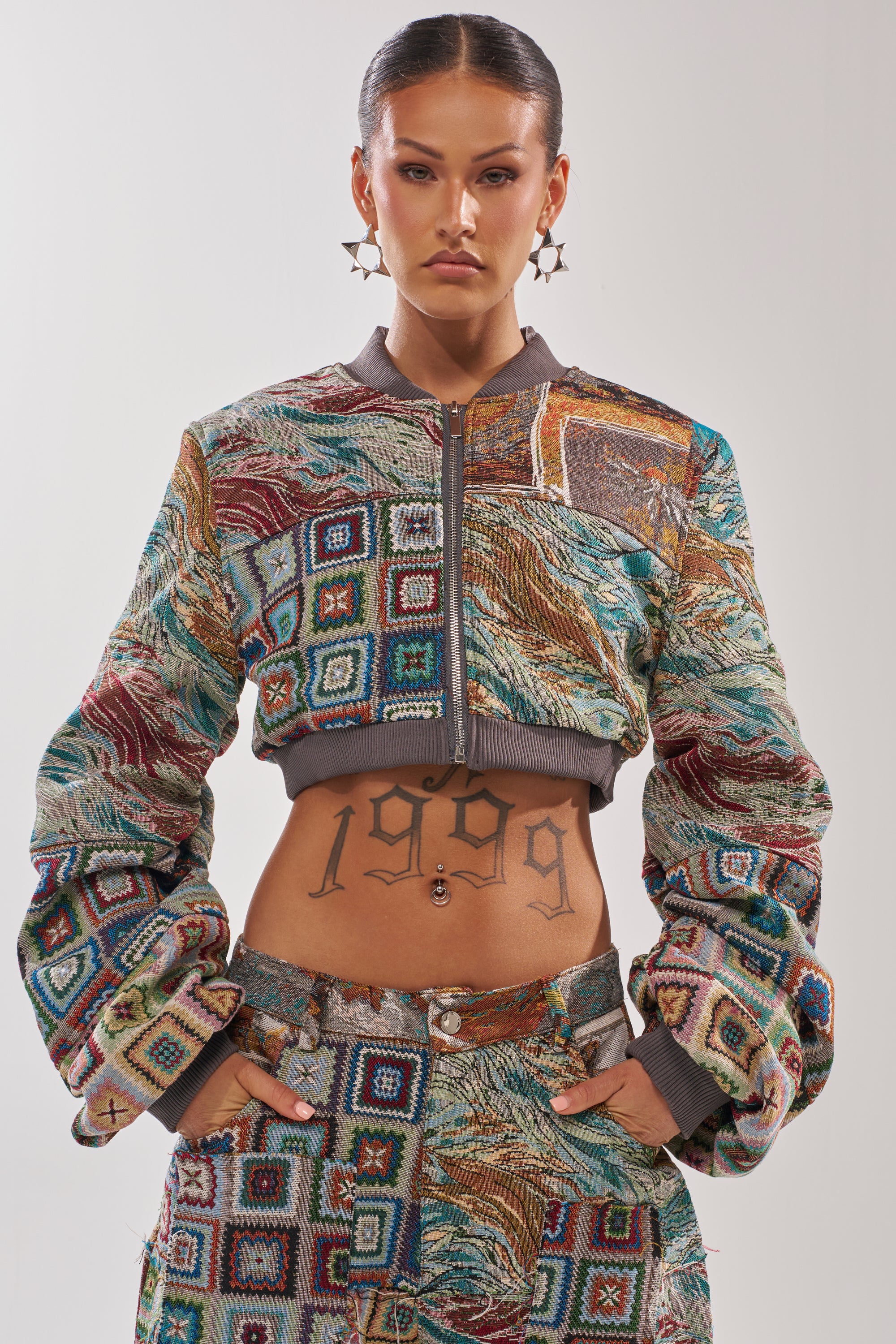 A woman exudes confidence in the WILD CROPPED PATCHWORK BOMBER and matching pants, featuring bold square and wave patterns. A "1999" tattoo, hoop earrings, and slicked-back hair complement the jacket’s vibrant tapestry texture. 0