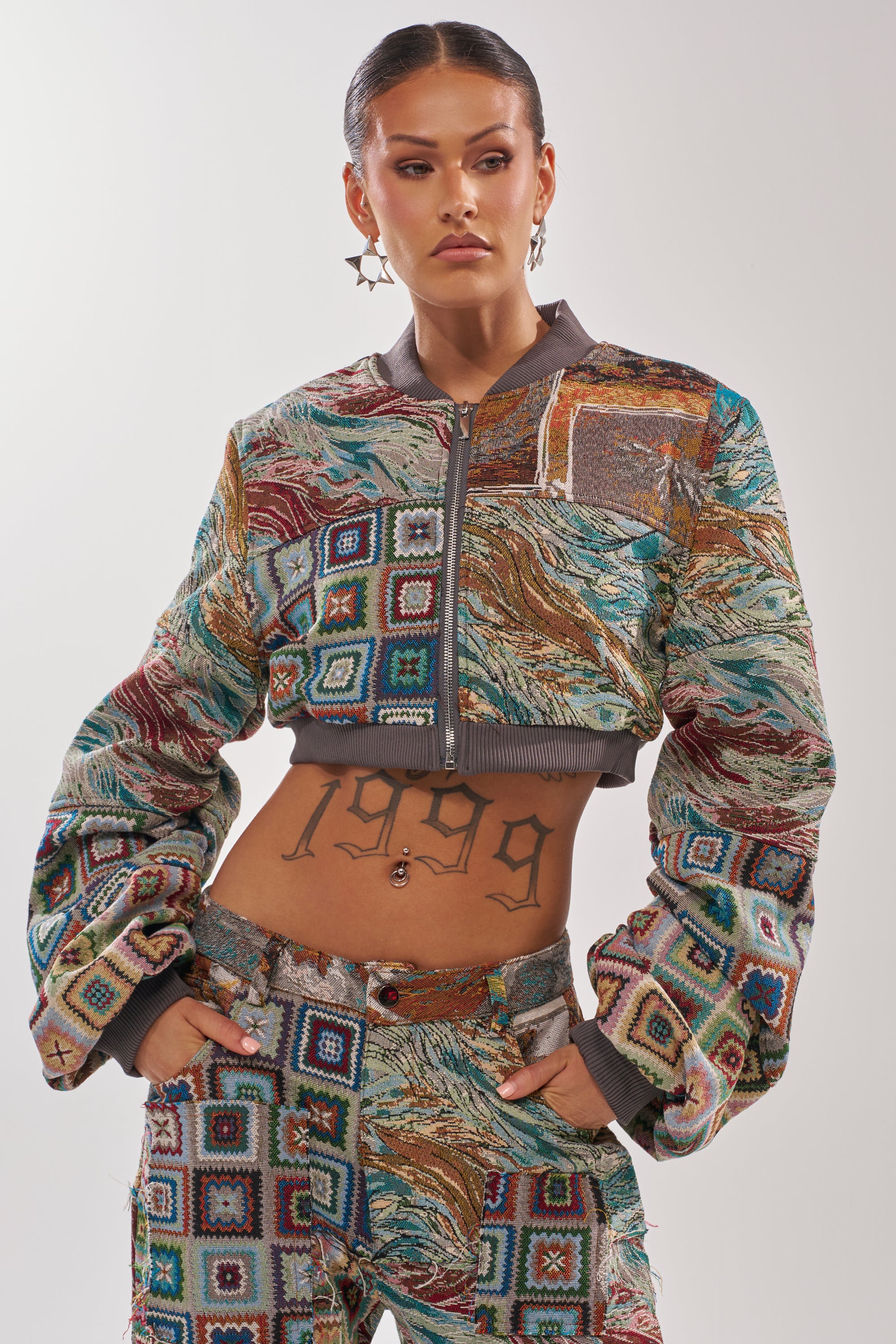 A woman poses confidently in the WILD CROPPED PATCHWORK BOMBER and matching pants. She sports star-shaped earrings, sleek hair, and a "1999" tattoo, highlighting the outfit’s bold textures and vibrant patchwork designs. 1