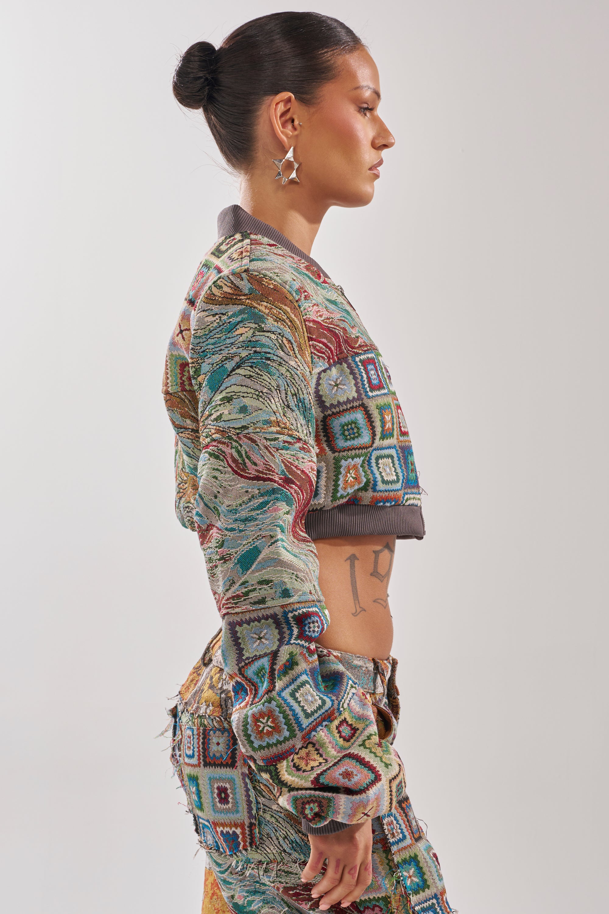 A woman with dark hair in a bun models the WILD CROPPED PATCHWORK BOMBER featuring geometric patterns. She stands sideways, revealing a side tattoo and star earrings, set against a plain background. 2