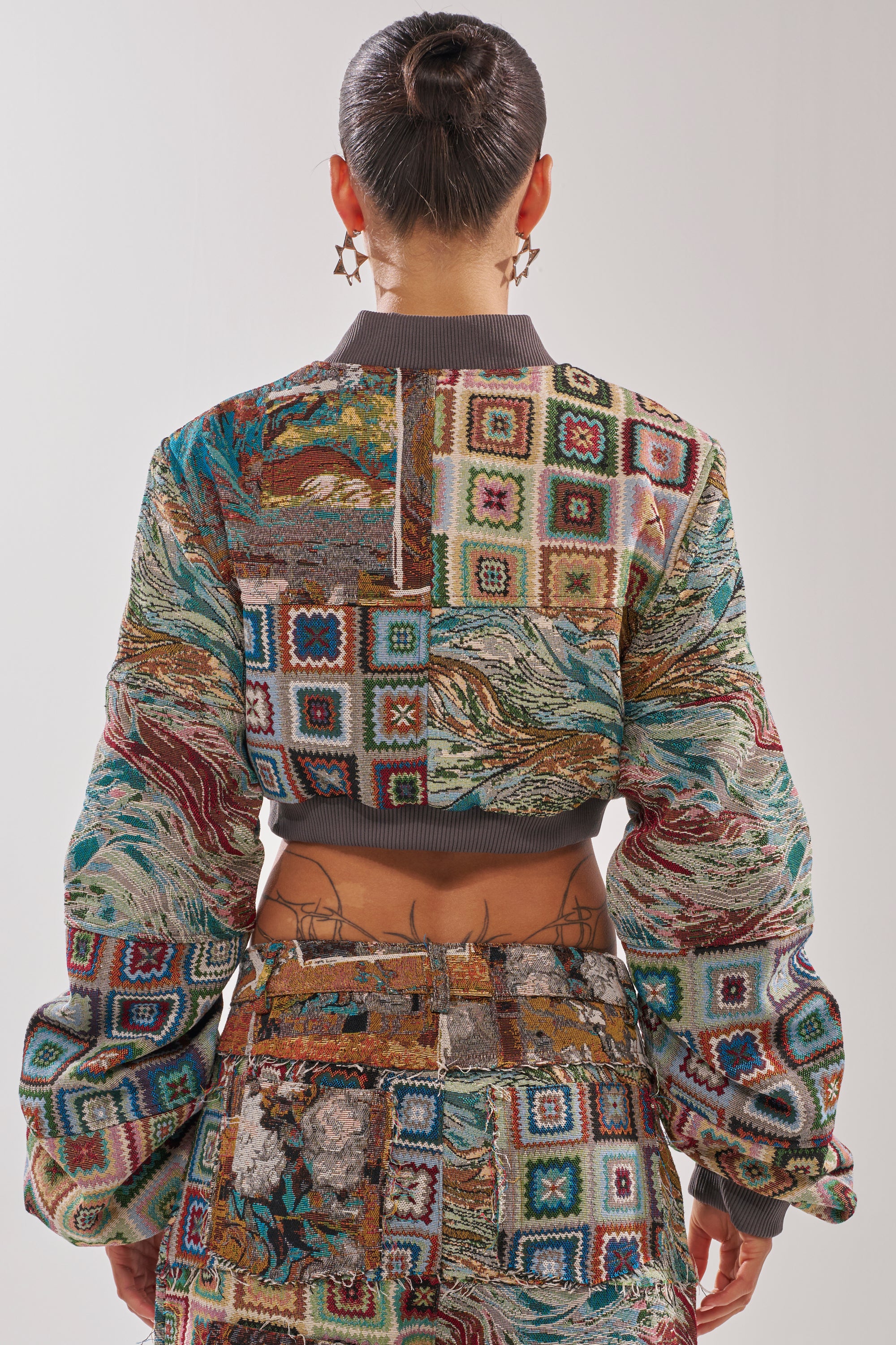 A person with dark hair in a sleek bun wears the WILD CROPPED PATCHWORK BOMBER and matching pants with geometric and abstract designs. Seen from the back, they display lower-back tattoos and wear star-shaped earrings. 3