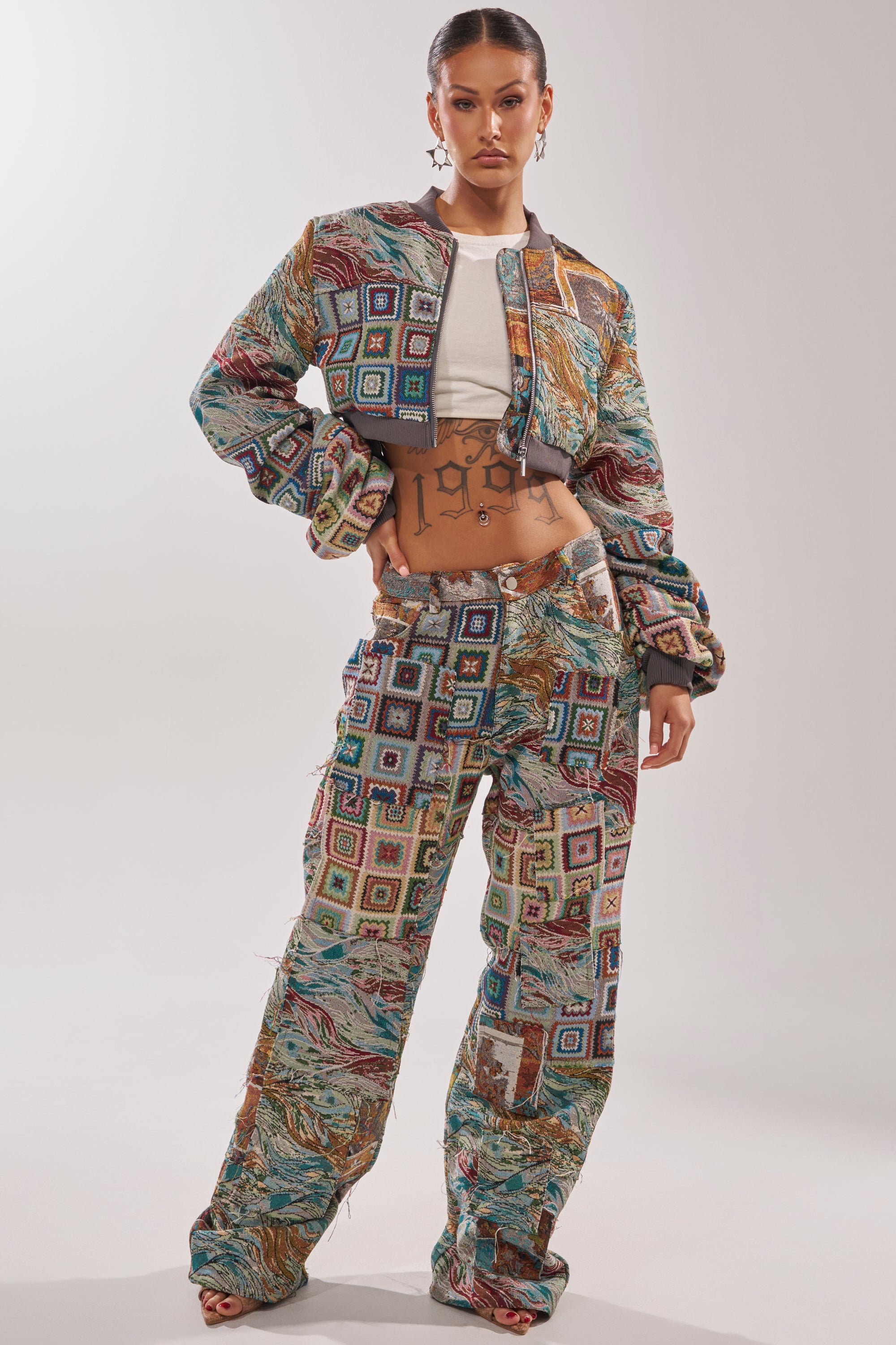 A woman poses confidently in the WILD CROPPED PATCHWORK BOMBER over a white crop top and wide-leg pants, showcasing hoop earrings and a bold “1996” tattoo on her abdomen. 4