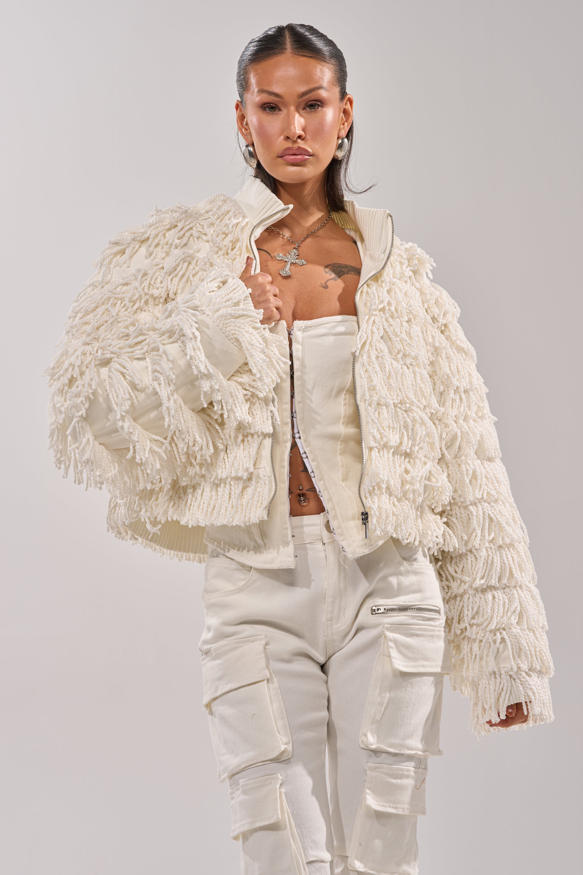 A woman stands confidently in the FREYA FRINGE BOMBER, a textured off-white jacket, styled over a white corset top and white cargo pants with zipper pockets against a plain background.