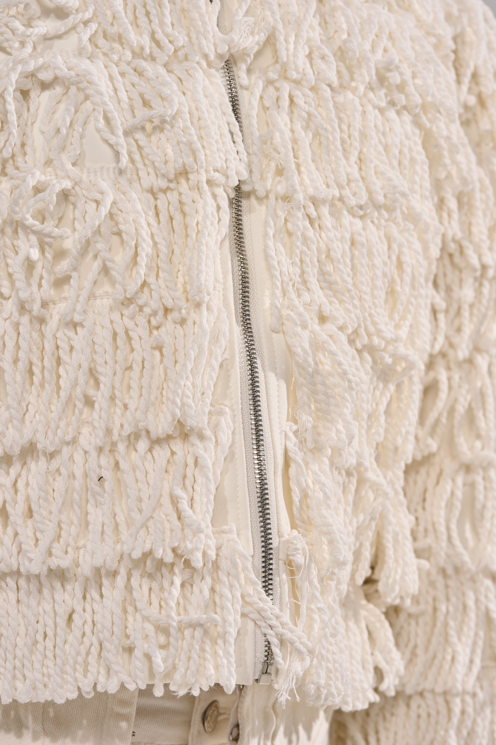 Close-up of the FREYA FRINGE BOMBER, a cream-colored jacket with a shaggy, looped yarn fringe and visible silver zipper, styled over matching pants.