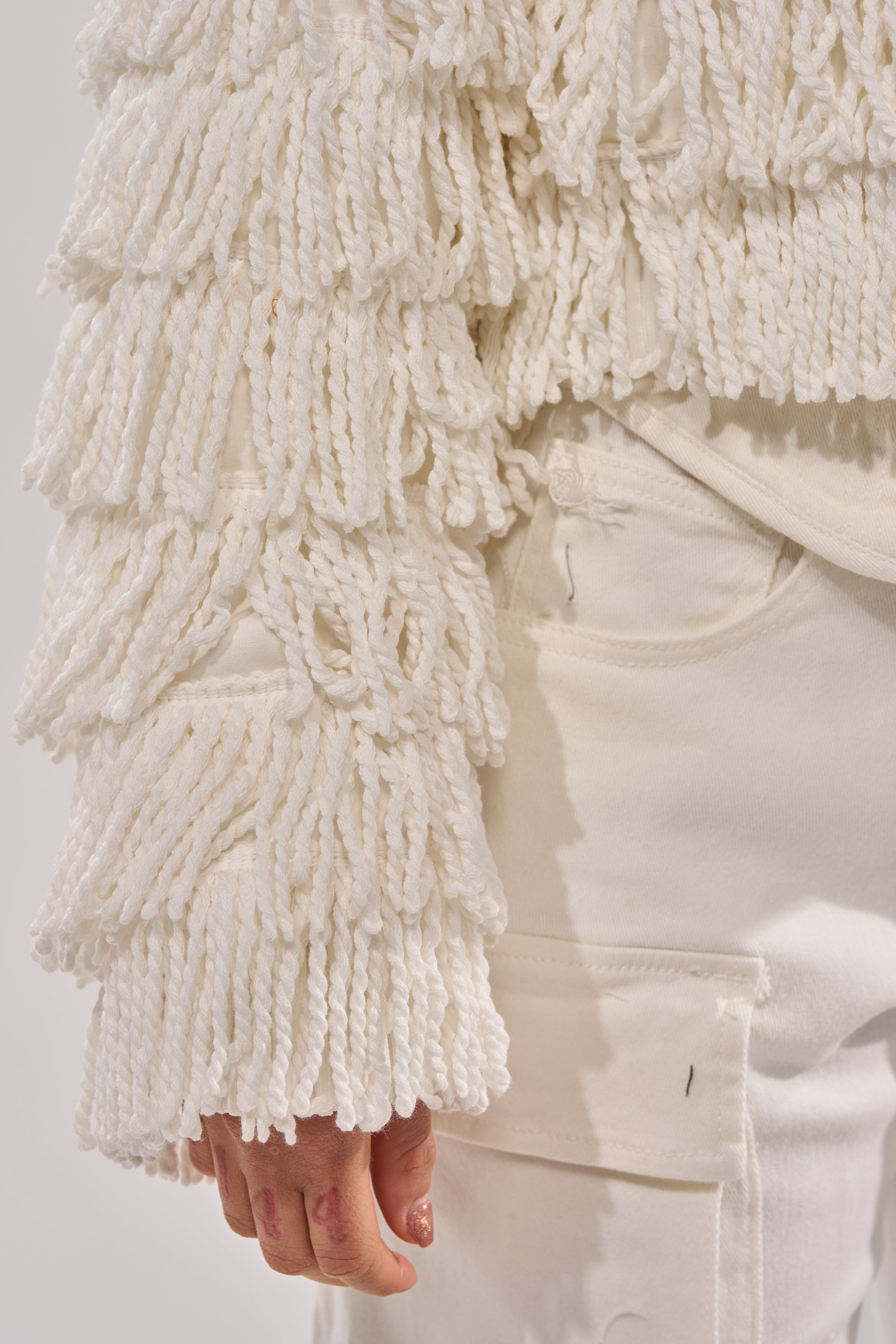A close-up of a person wearing cream pants and the FREYA FRINGE BOMBER, highlighting its shaggy textured, looped sleeve and pocket detail.