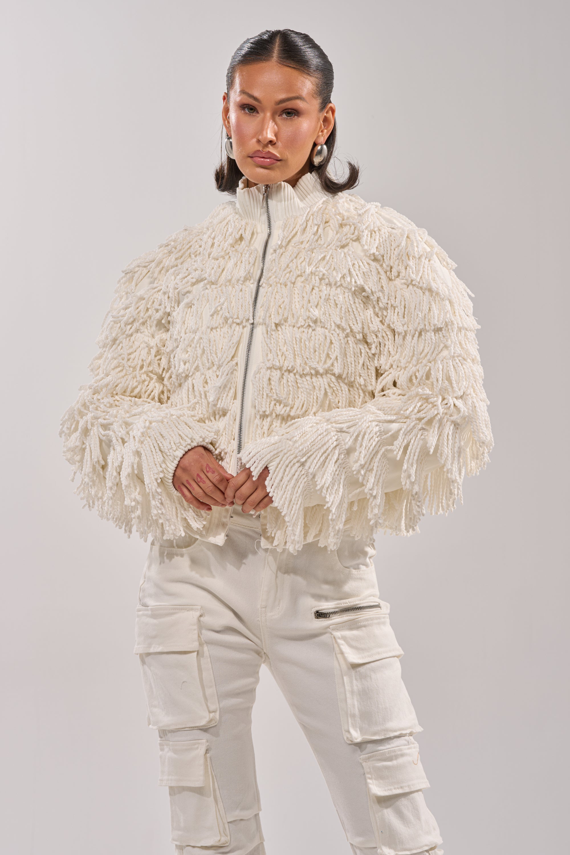 A woman confidently poses against a plain background, wearing the FREYA FRINGE BOMBER in cream with matching cargo pants and her hands clasped.