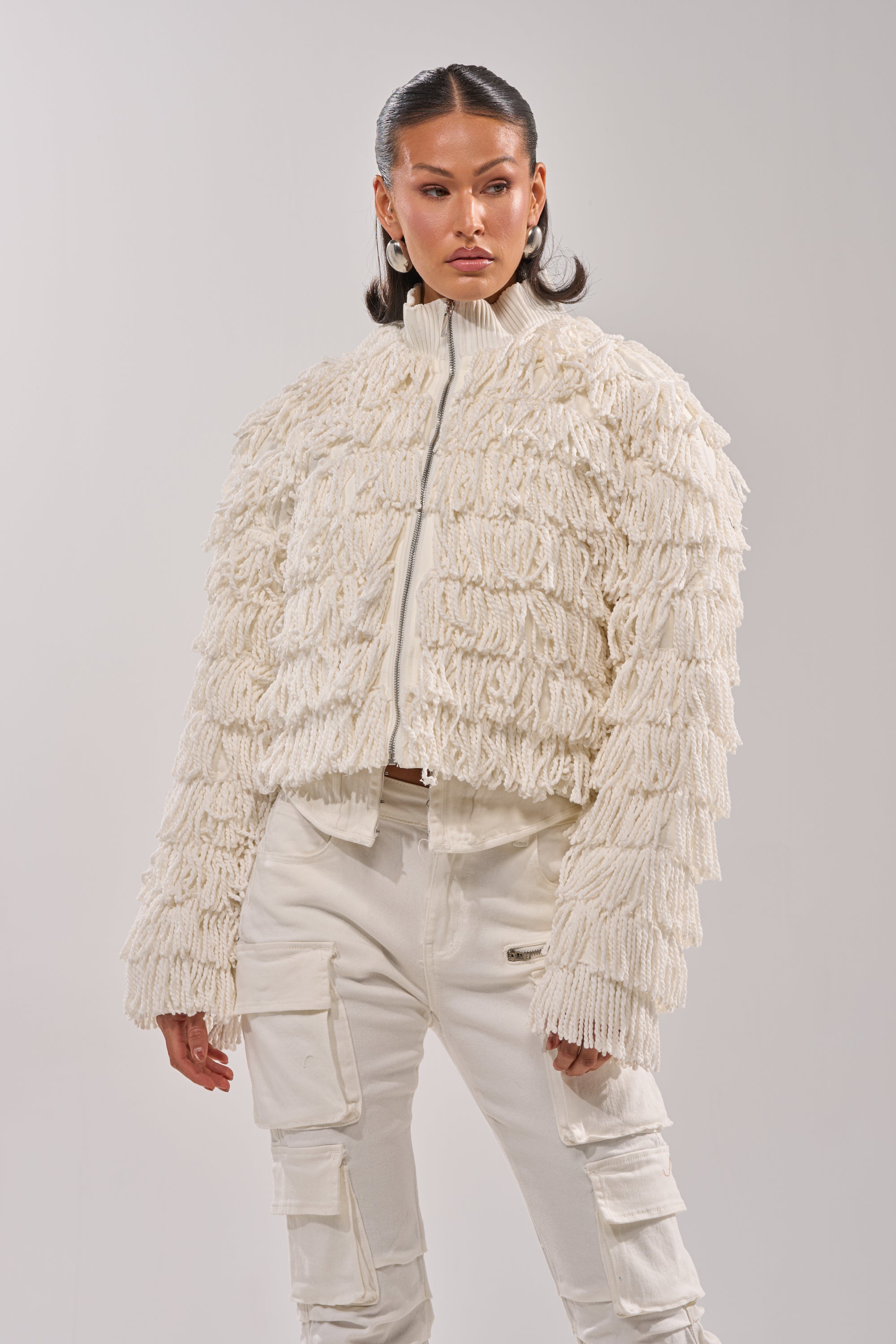 A woman confidently stands against a plain light background, wearing the FREYA FRINGE BOMBER—an off-white, textured jacket with a shaggy fringe finish—paired with matching white cargo pants.