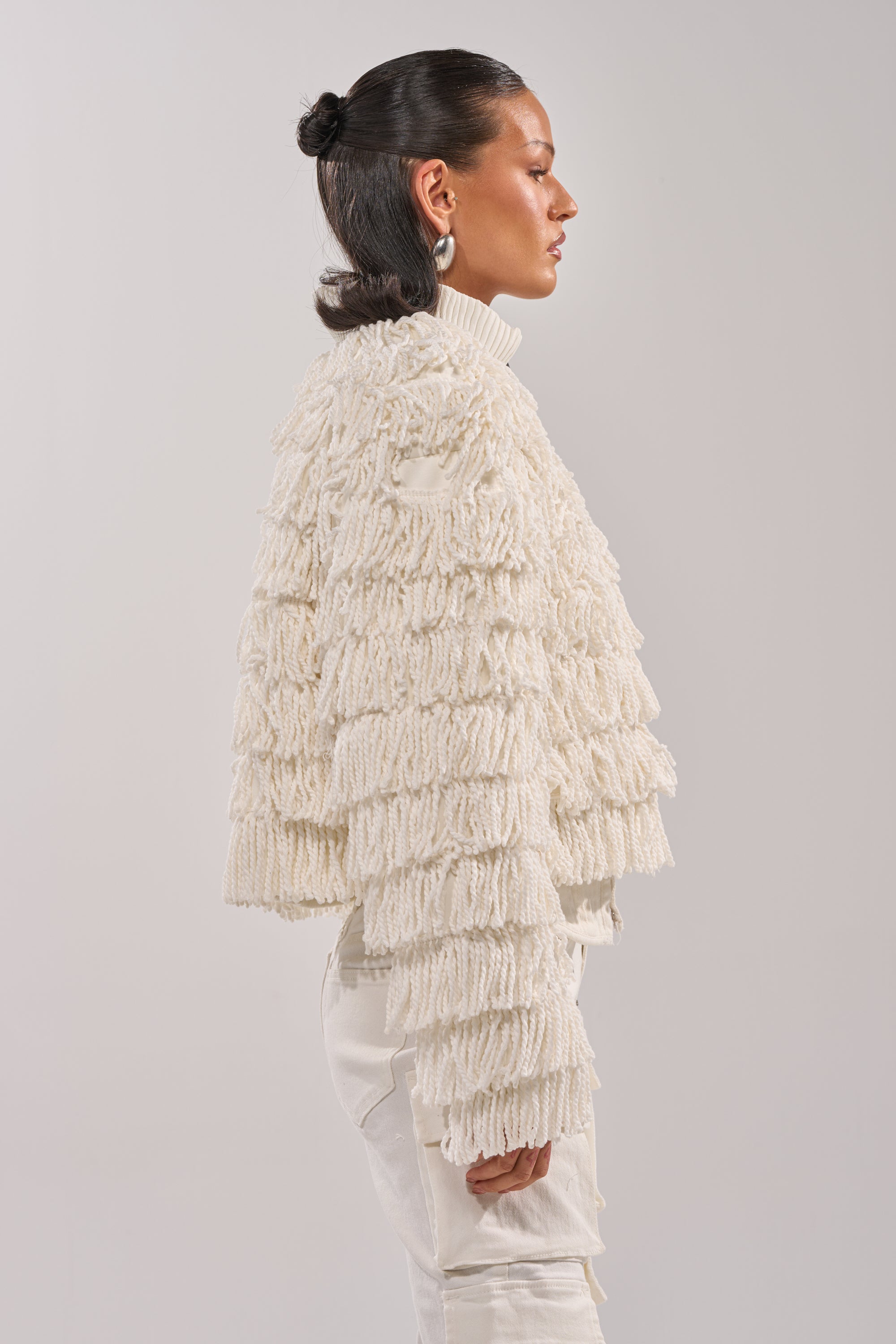 A person with dark, slicked-back hair in a bun wears the FREYA FRINGE BOMBER, a textured off-white cropped jacket, paired with a white turtleneck and pants, standing in profile against a plain light background.